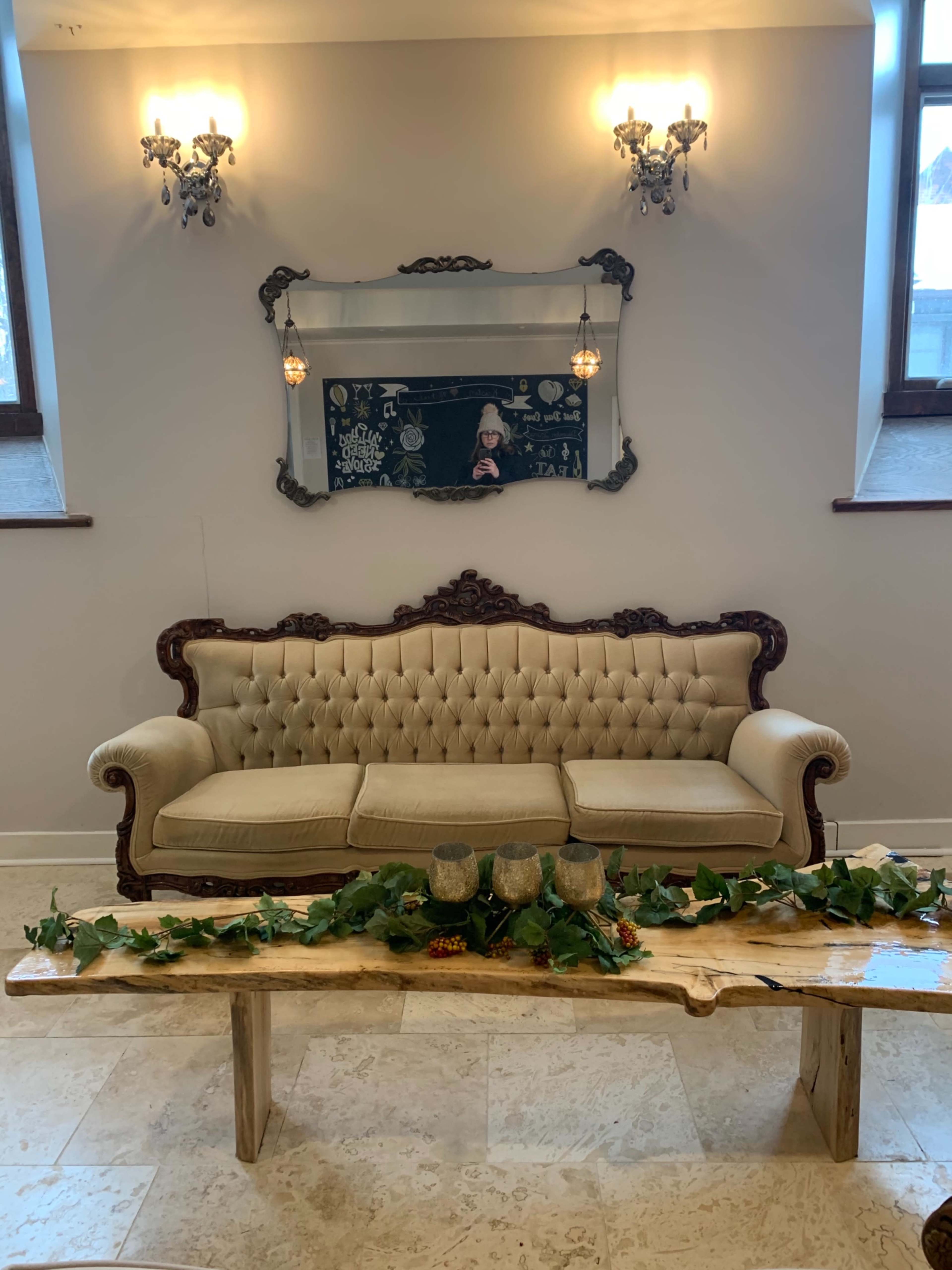 A vintage tufted sofa is positioned against a neutral wall, accompanied by a rustic wooden table decorated with greenery and two decorative glasses, with a framed mirror and wall sconces above.