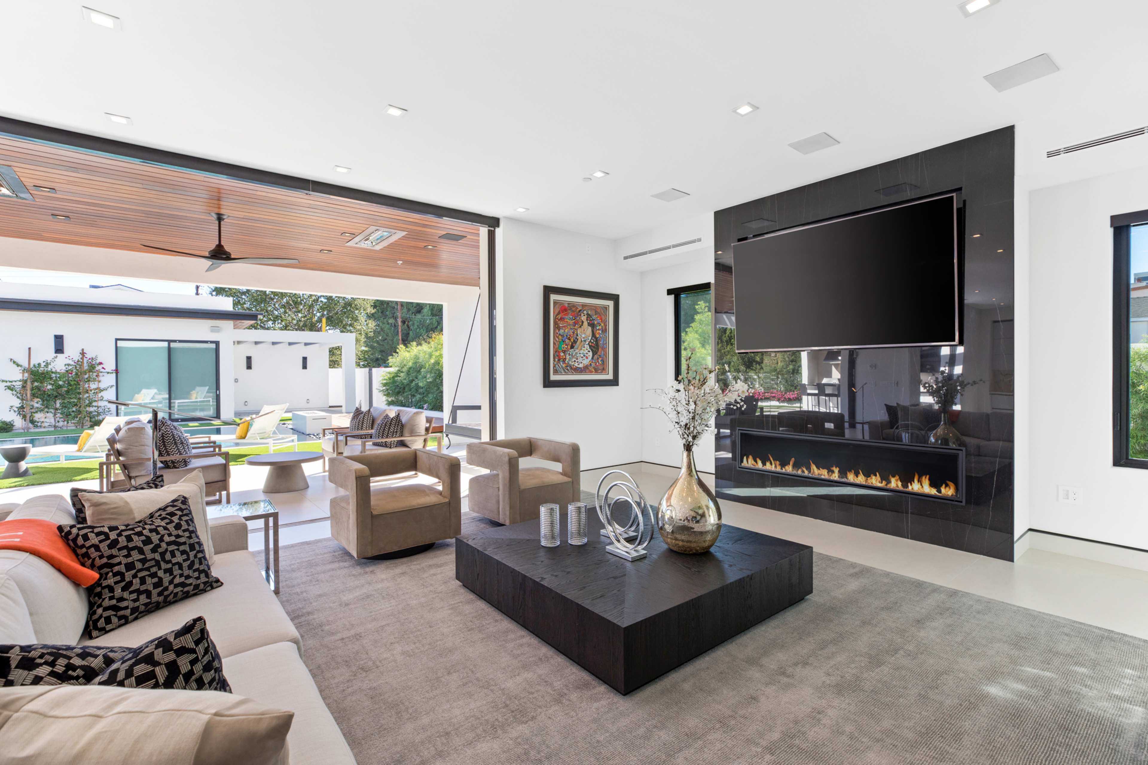 The image shows a modern living room with a large flat-screen TV mounted on a black wall, a coffee table in the center, and expansive windows that open to an outdoor seating area.
