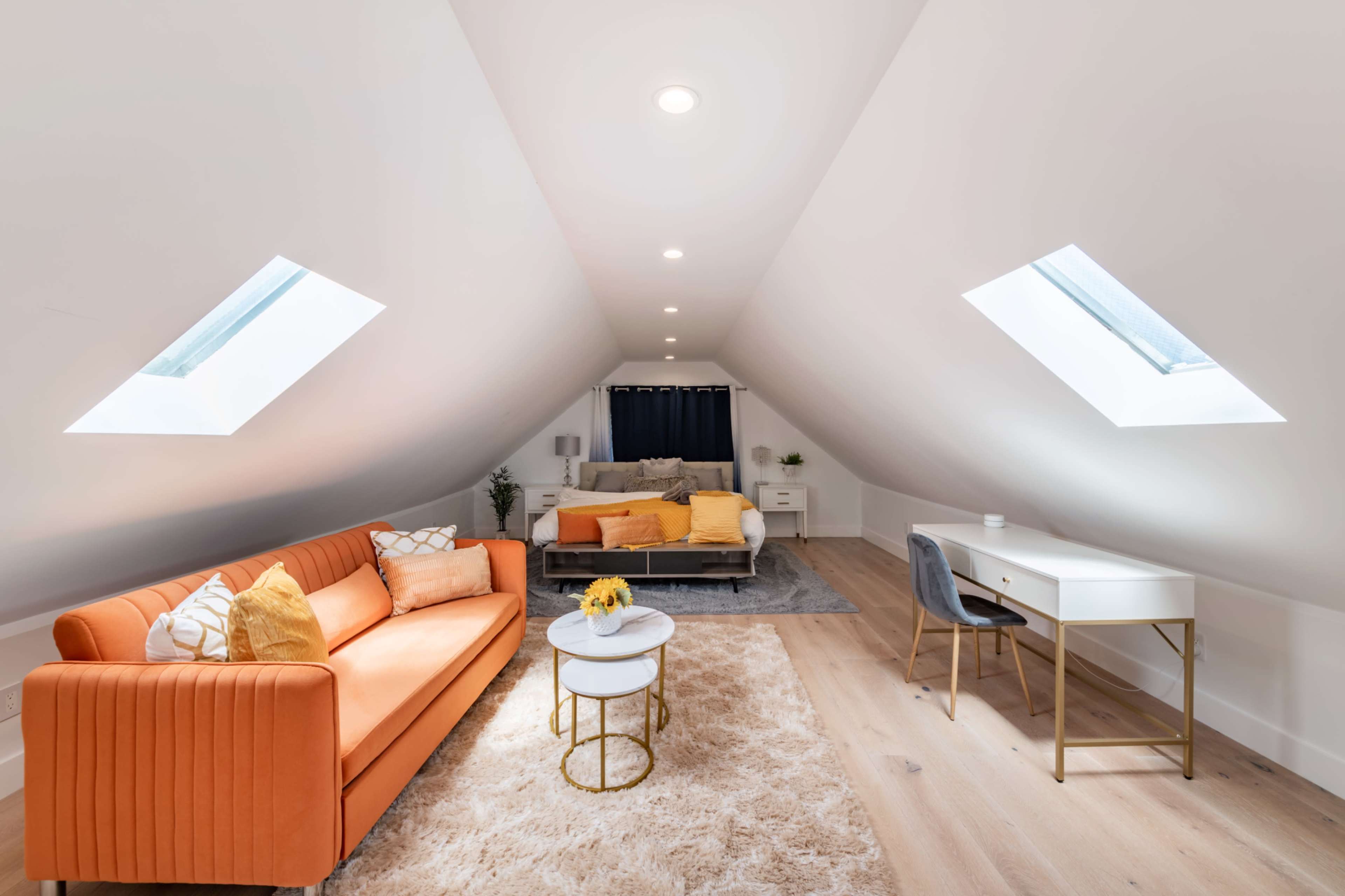 The image shows a bright attic space featuring a cozy orange sofa, a small coffee table, a bed with colorful linens, and a desk under slanted skylights.