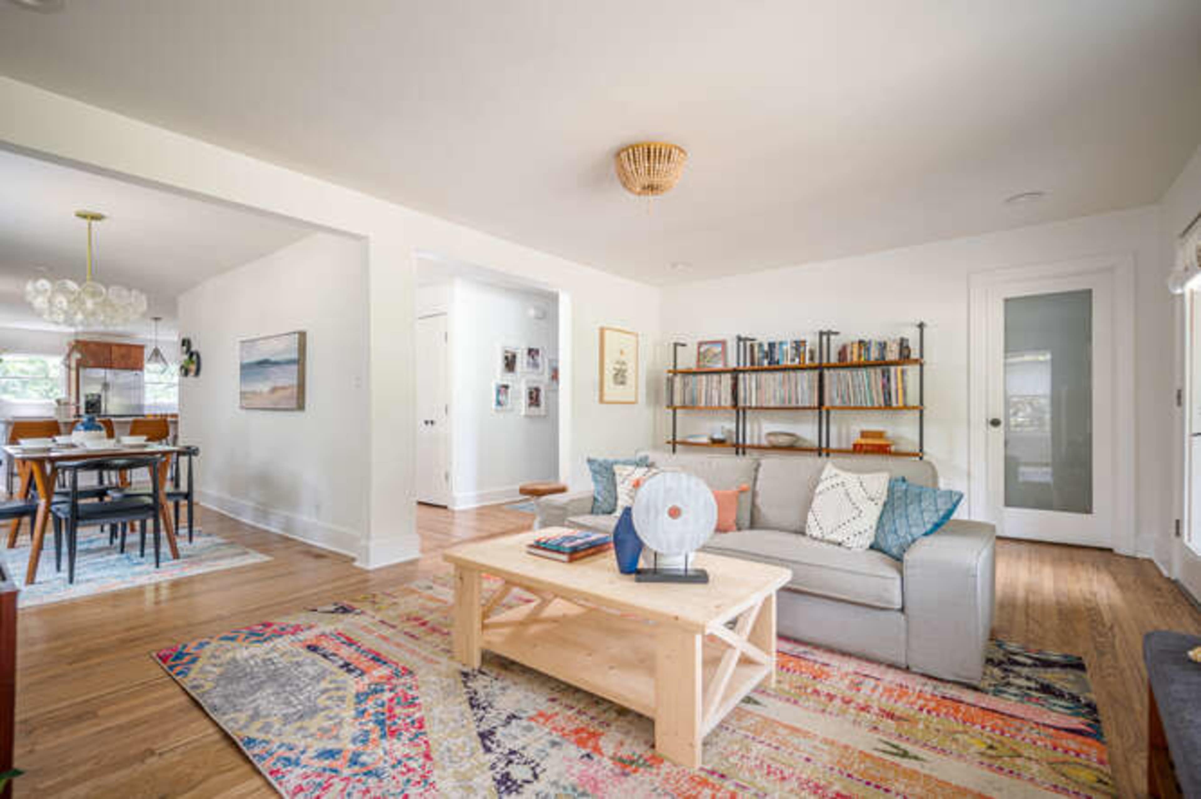 A cozy living room features a light gray sofa, a wooden coffee table, and a colorful area rug, with a bookshelf in the background and a dining area visible through an adjacent doorway.
