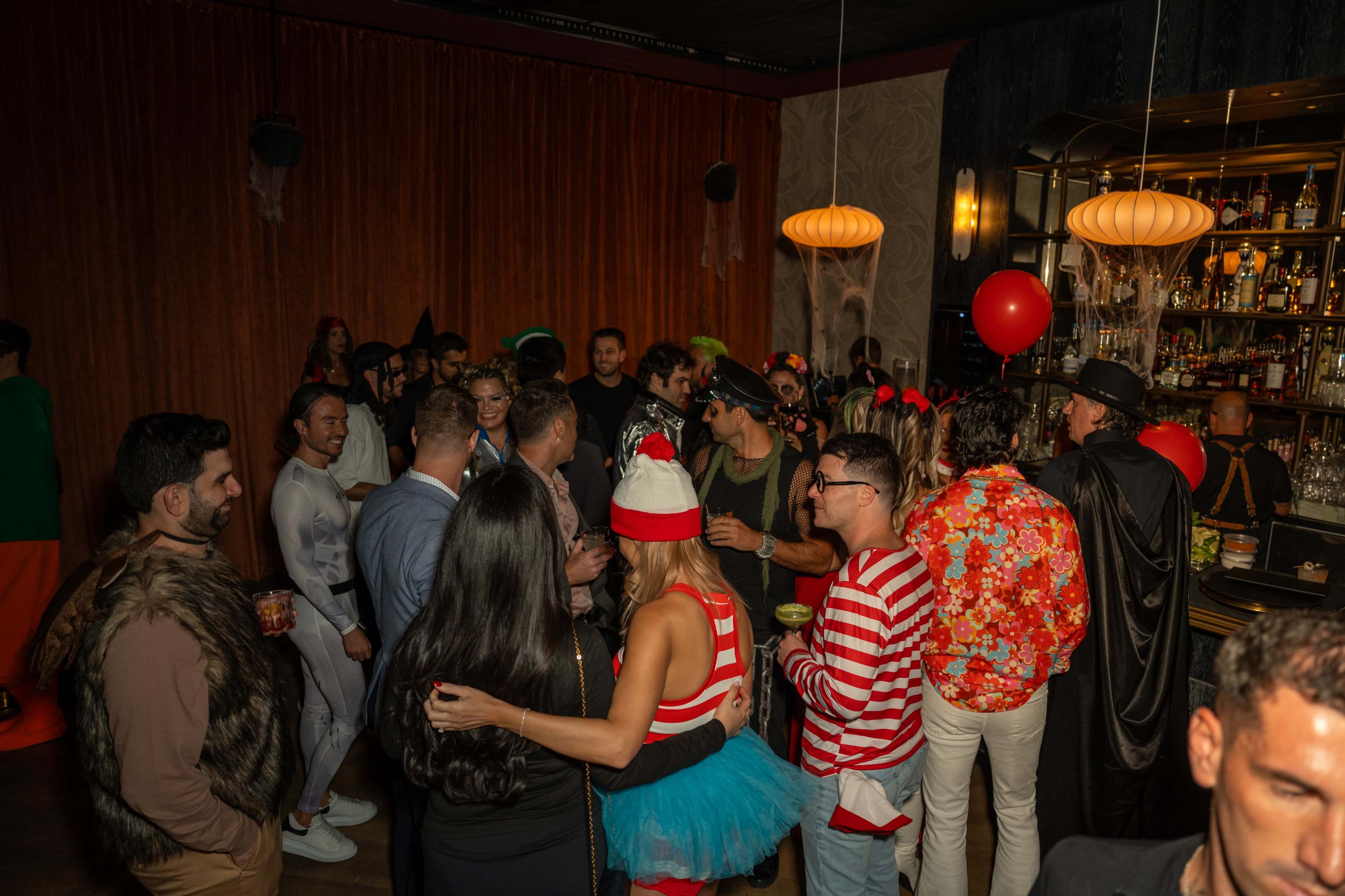 A group of people are gathered in a lively bar, dressed in various costumes and enjoying drinks during a festive event.