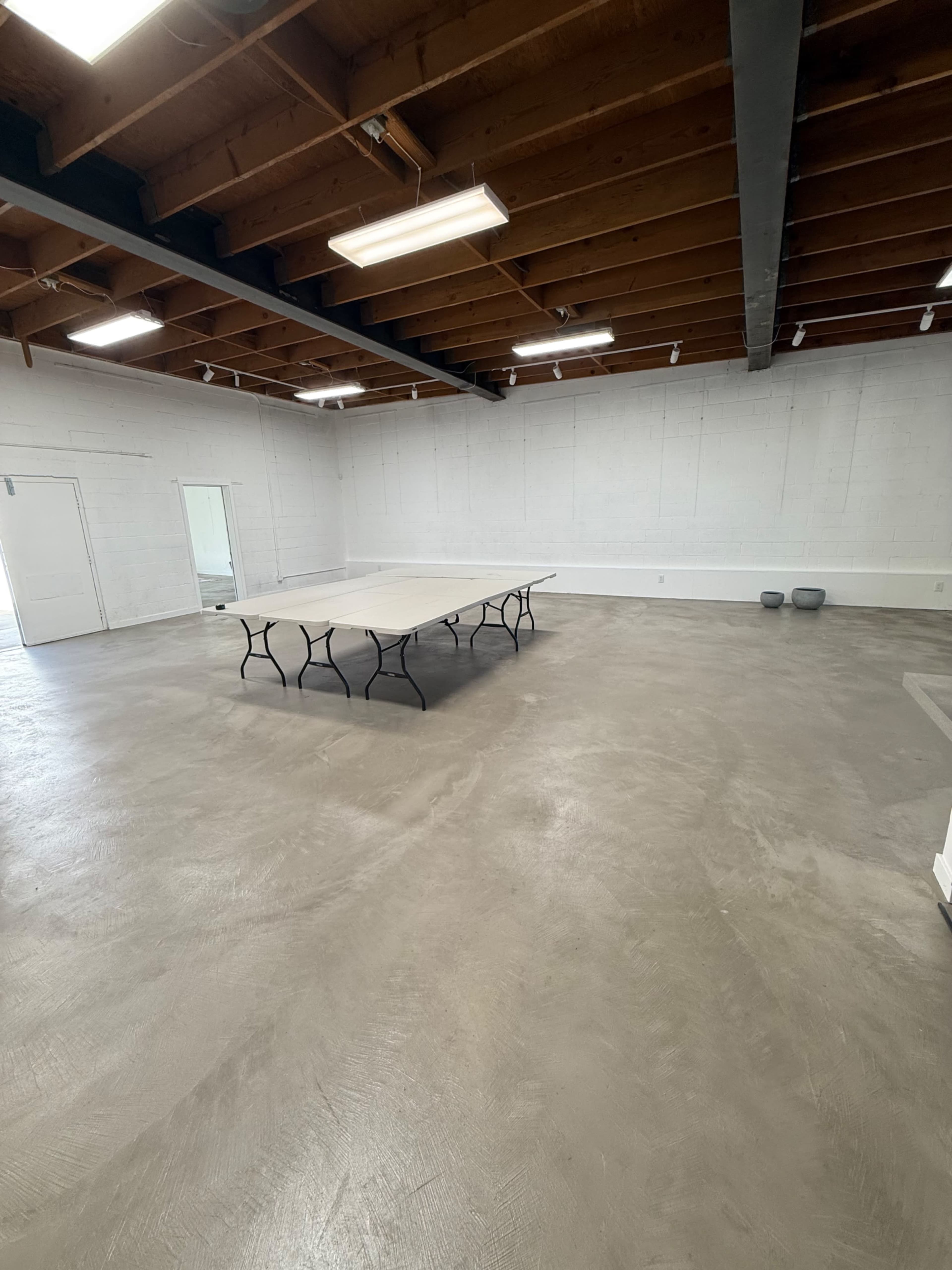 The image shows a spacious, empty room with a flat concrete floor, a long table in the center, and exposed wooden beams on the ceiling.