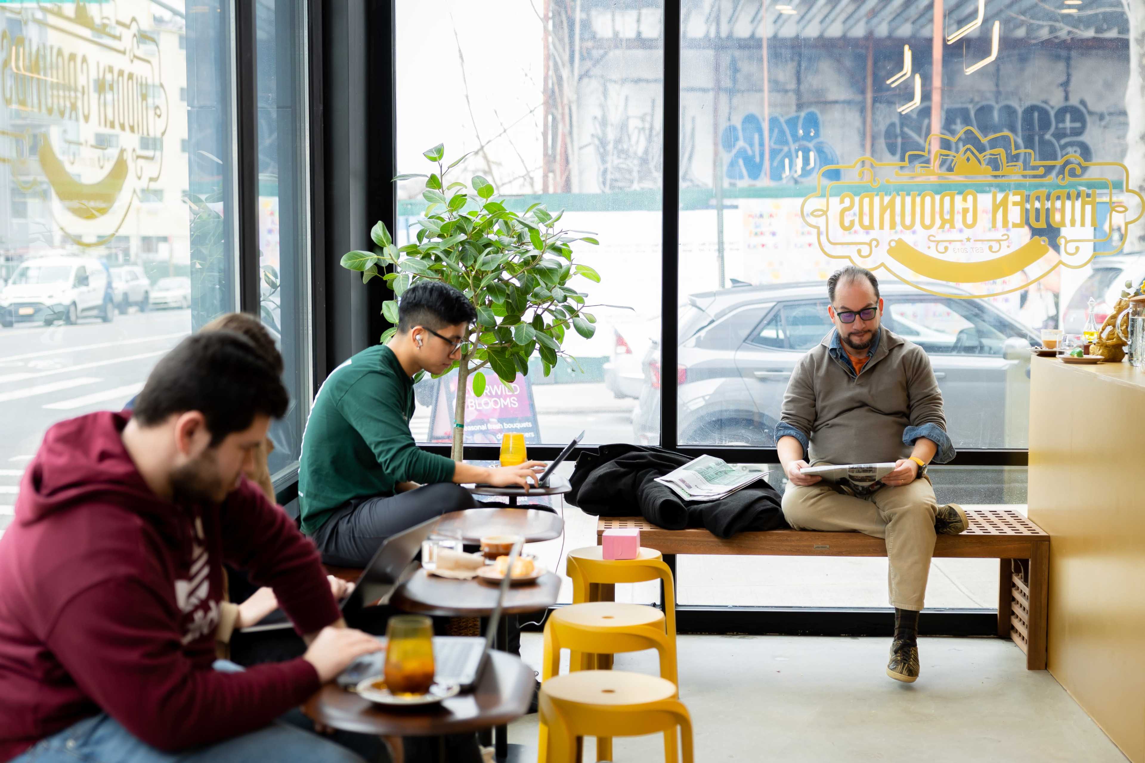 A modern café features two customers working on laptops while a third reads a newspaper, all seated at a long table with a view of the street.