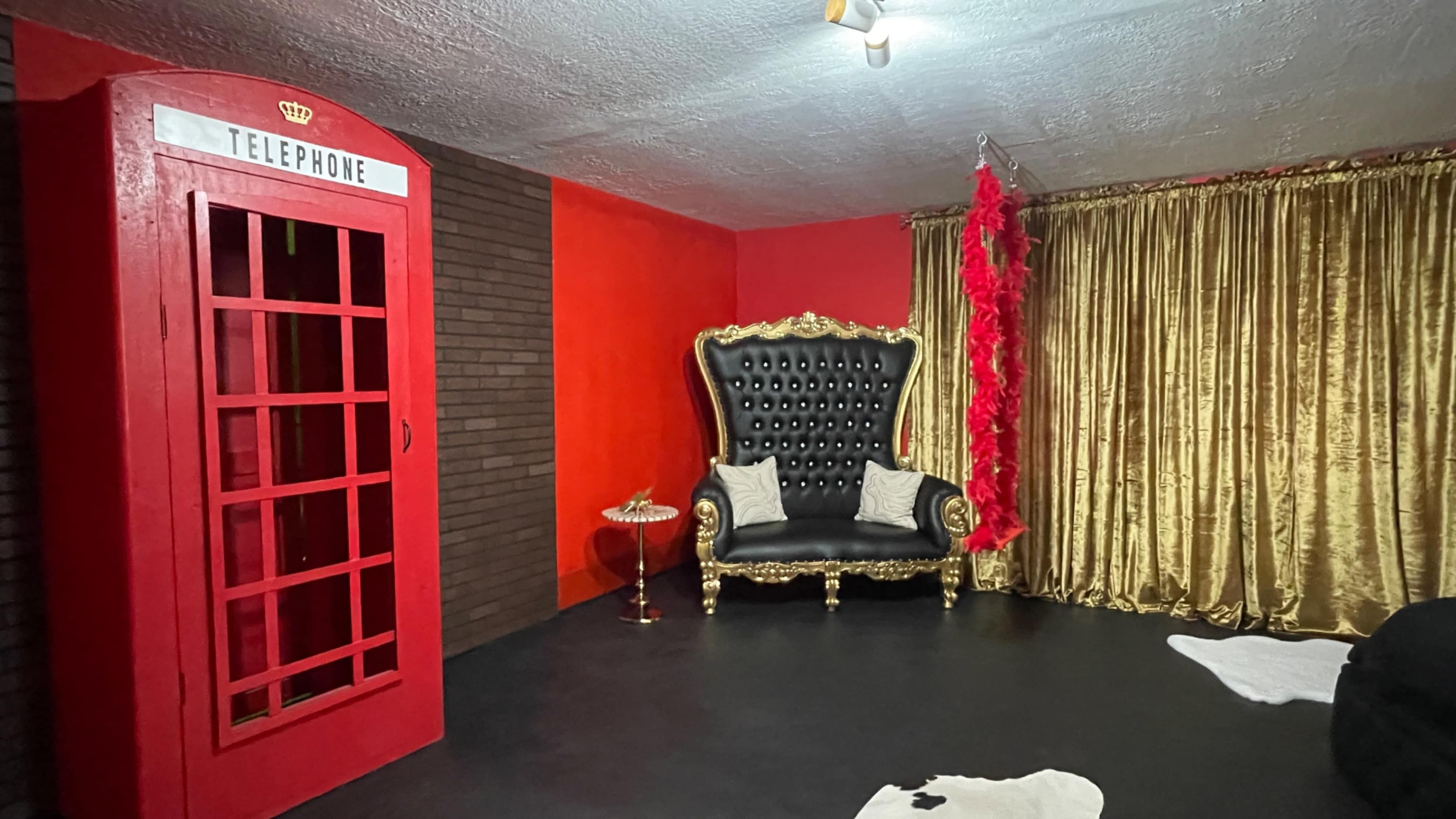 A room featuring a red telephone booth, a black upholstered sofa with gold accents, and gold curtains on a wall.