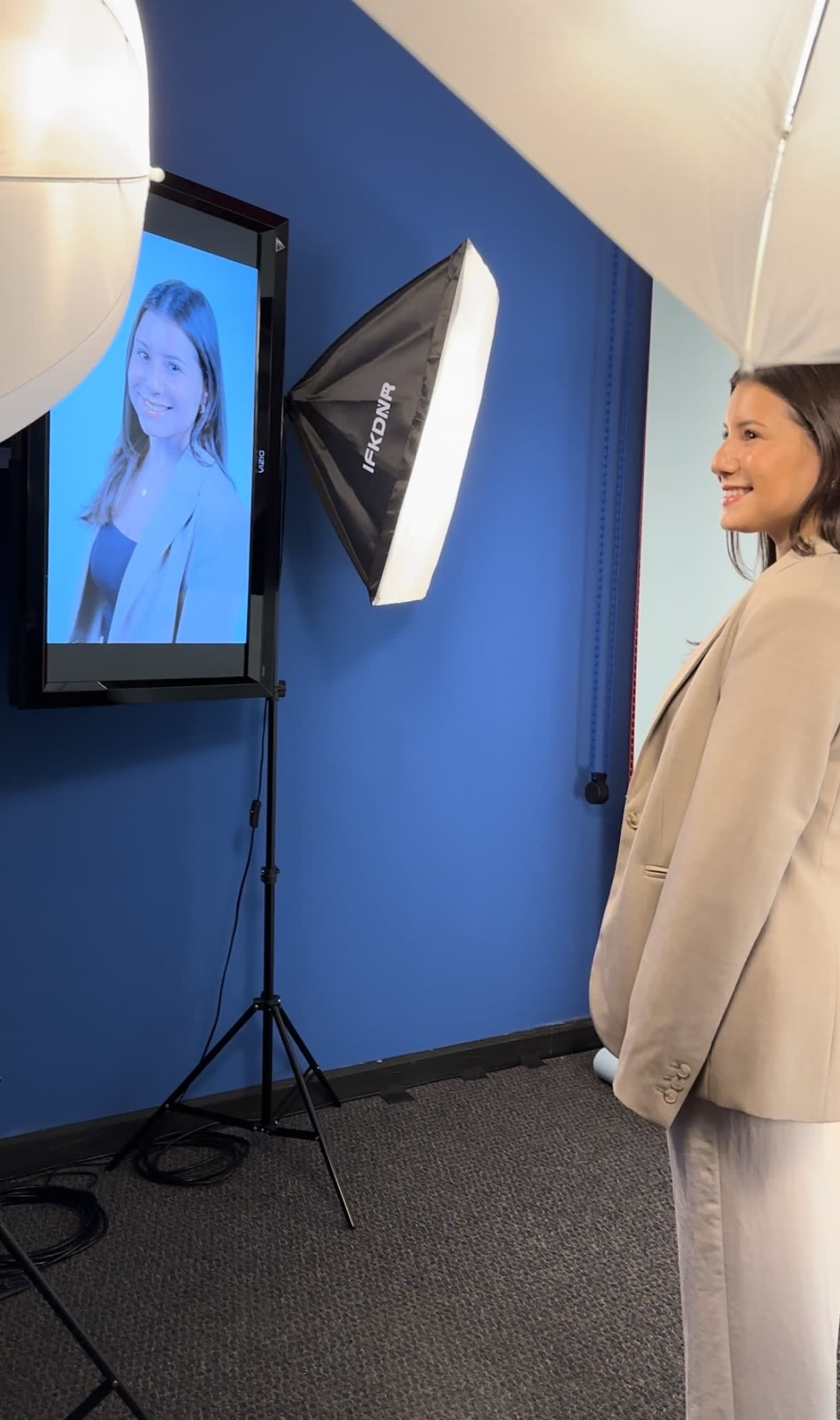 A woman stands in front of a camera setup, smiling while looking at her image displayed on a monitor.