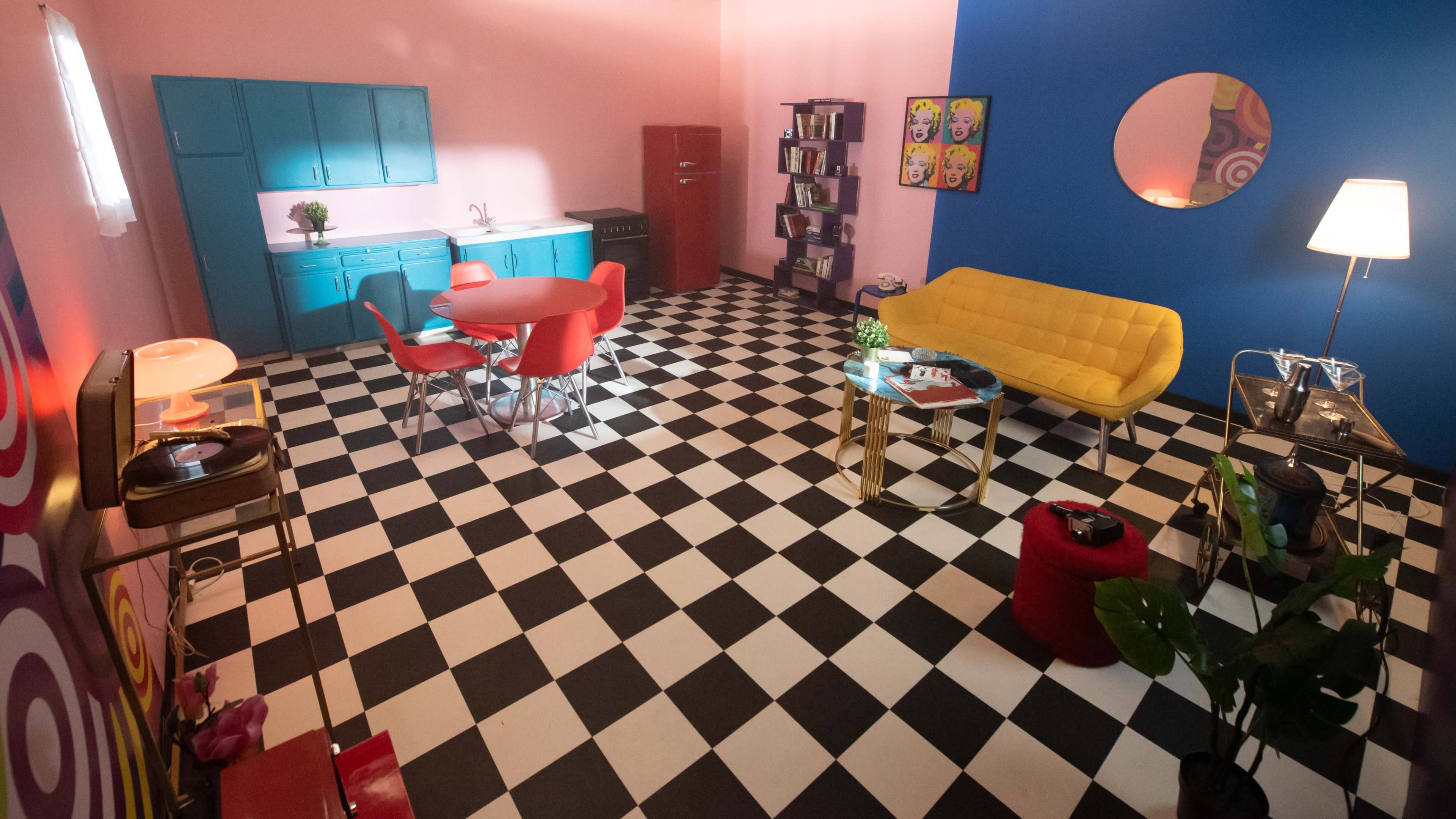 A brightly colored room with a checkered black-and-white floor featuring a blue accent wall, a yellow couch, red dining chairs, and a small kitchenette.