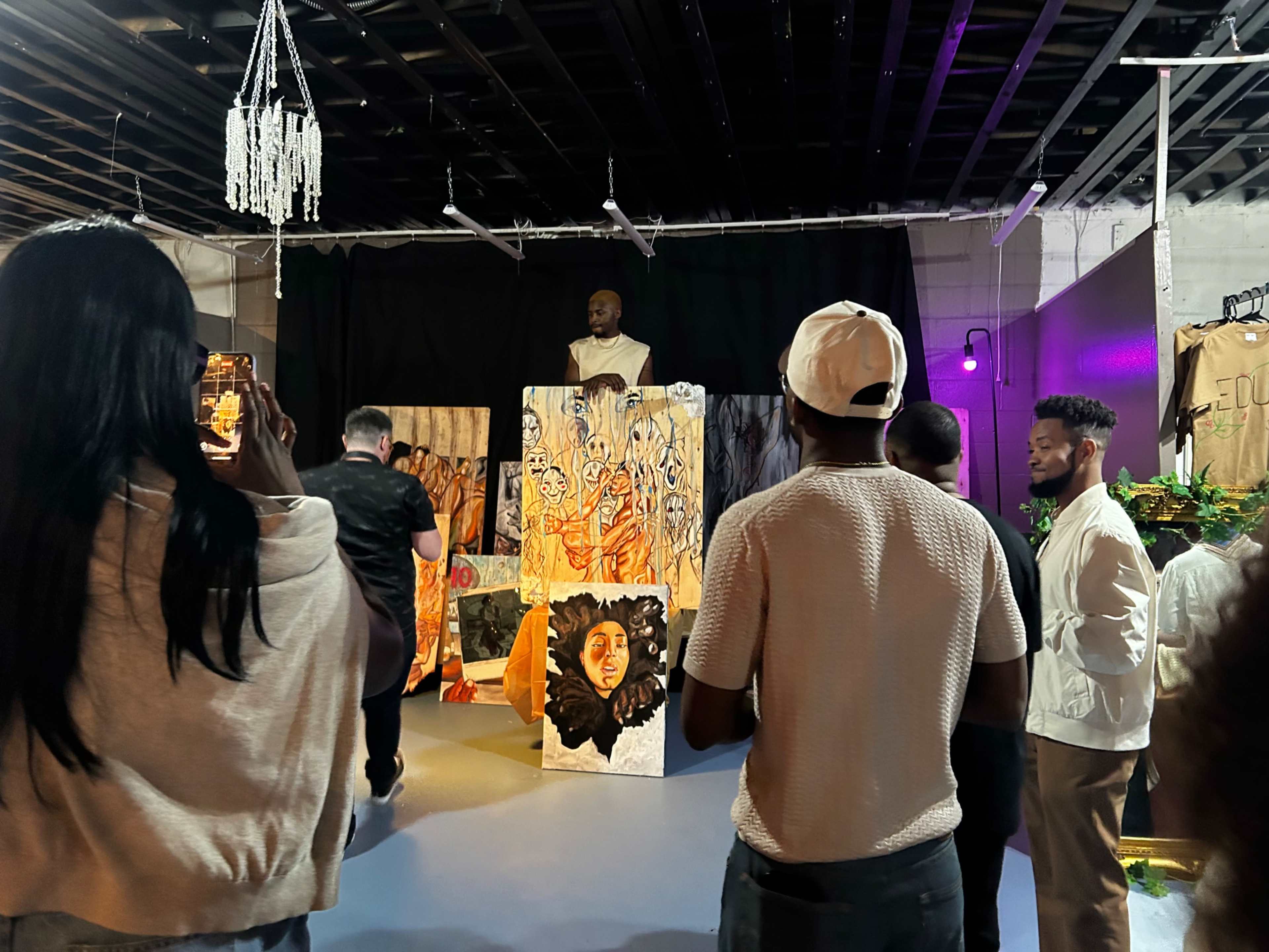 A crowd gathers around an artist standing on a pedestal amidst colorful artwork in a gallery setting.