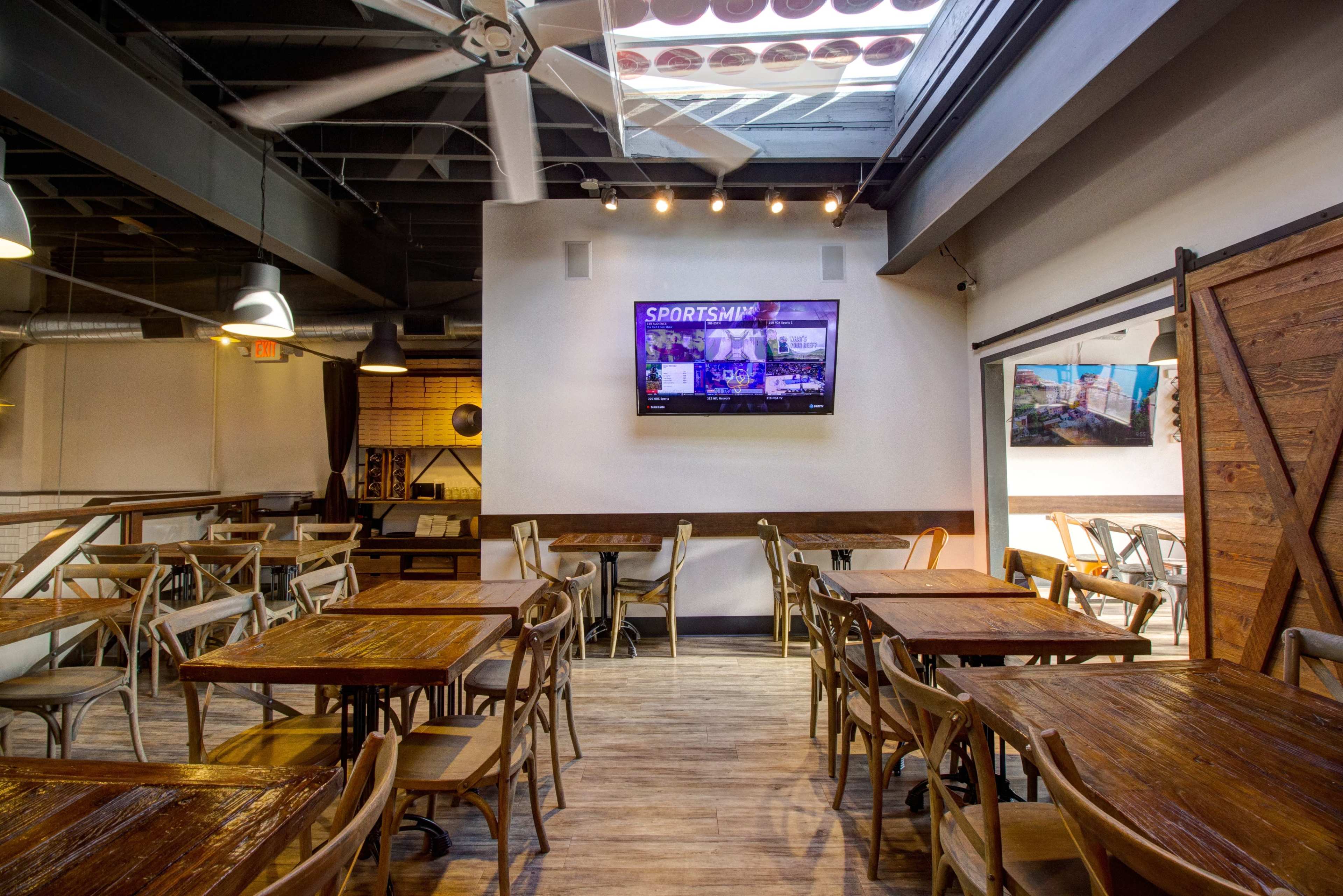 A spacious restaurant interior with wooden tables and chairs, featuring a large wall-mounted TV displaying sports content.
