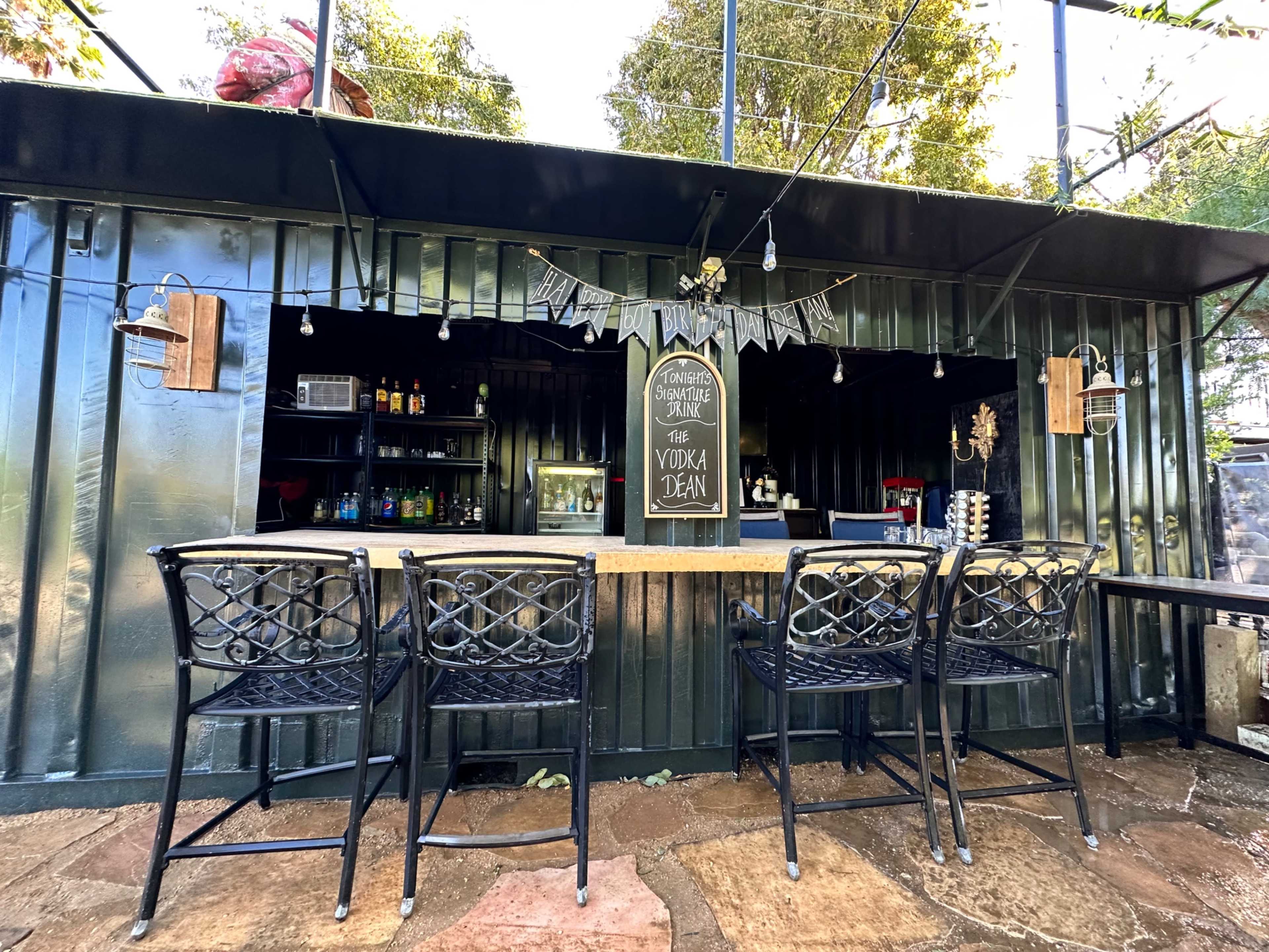 An outdoor bar area with a counter, several black metal stools, and string lights hanging above.
