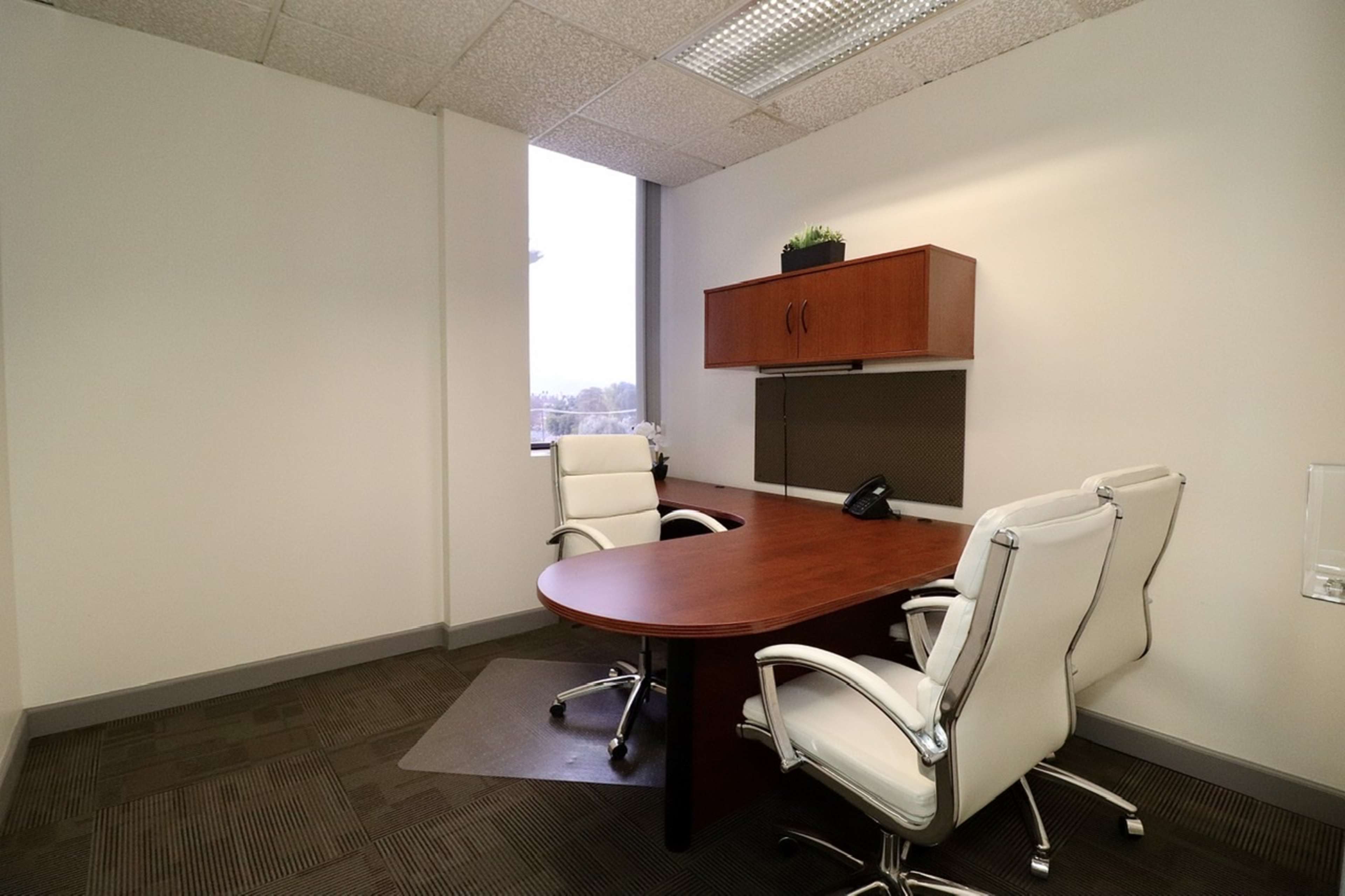 An empty office space featuring a round wooden table and two white swivel chairs, with a large window and a wooden cabinet mounted on the wall.