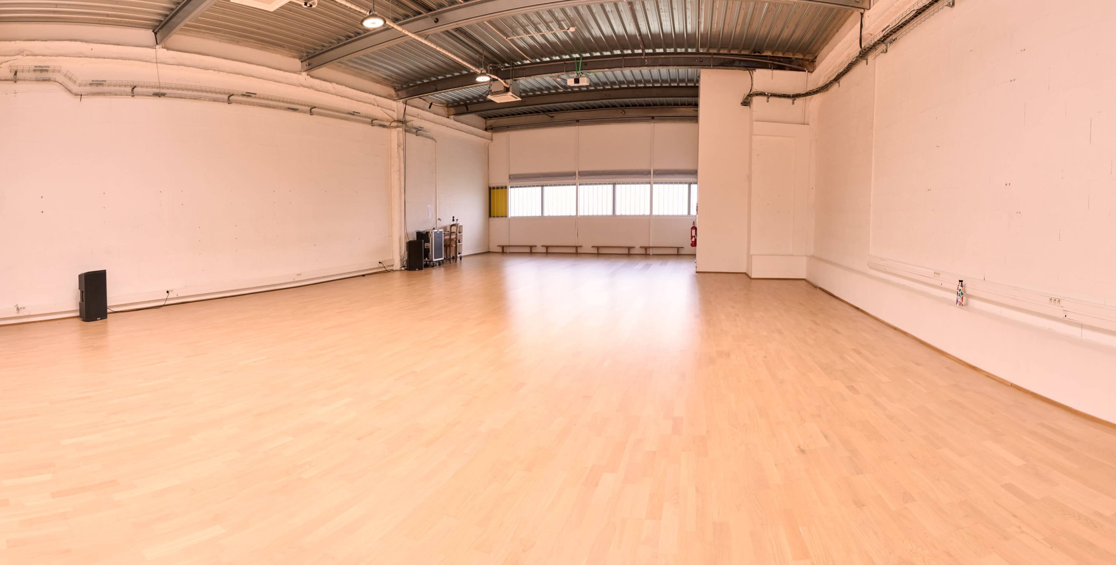 The image shows a spacious, empty room with wooden flooring and large windows providing natural light.