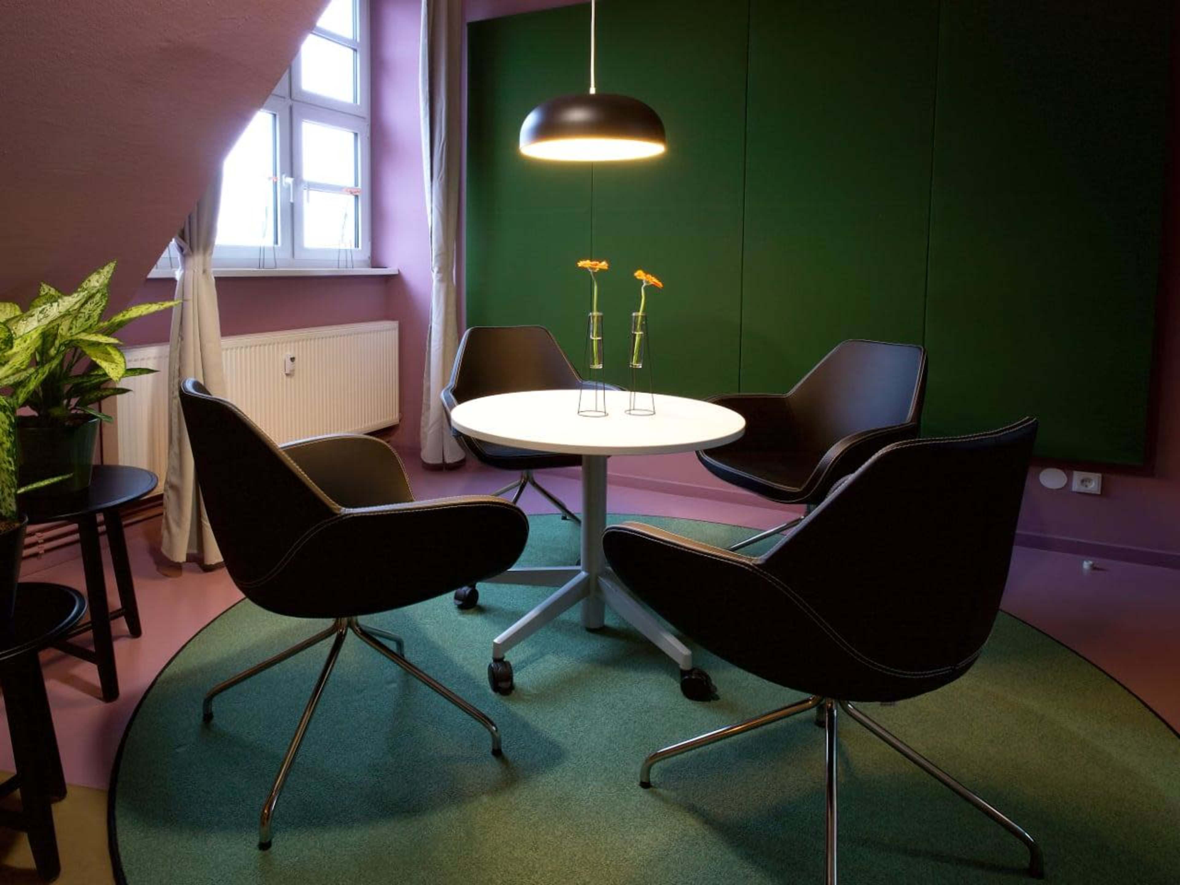 A small meeting room features a round table surrounded by four modern chairs, with a potted plant nearby and a large window letting in natural light.