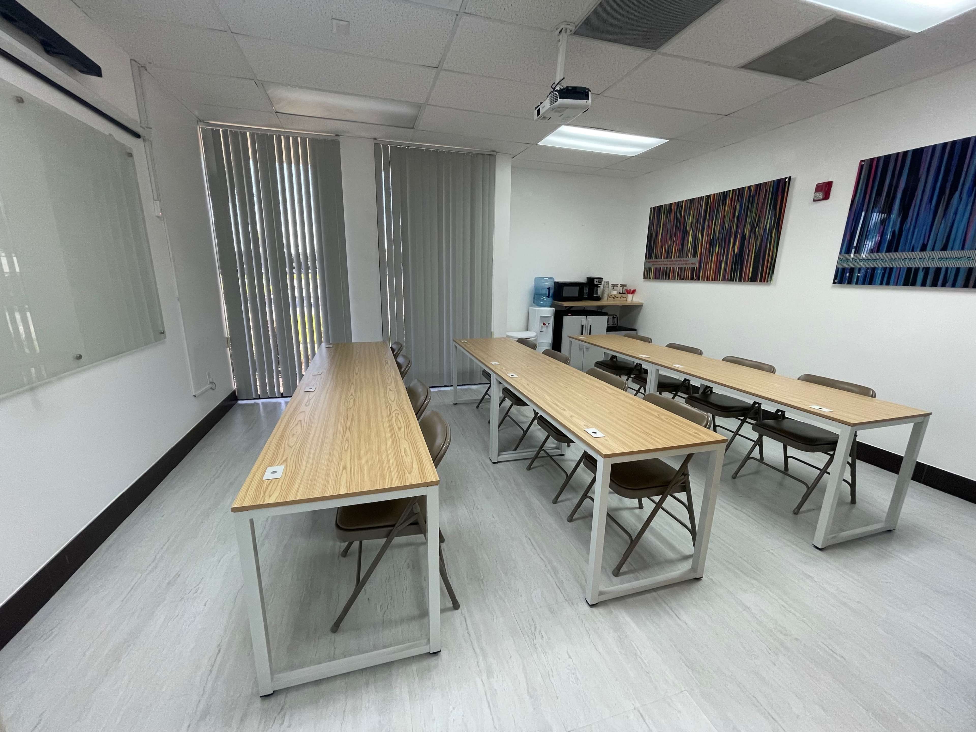 A classroom with two long tables arranged in an L-shape, several chairs, a projector mounted on the ceiling, and artwork on the walls.