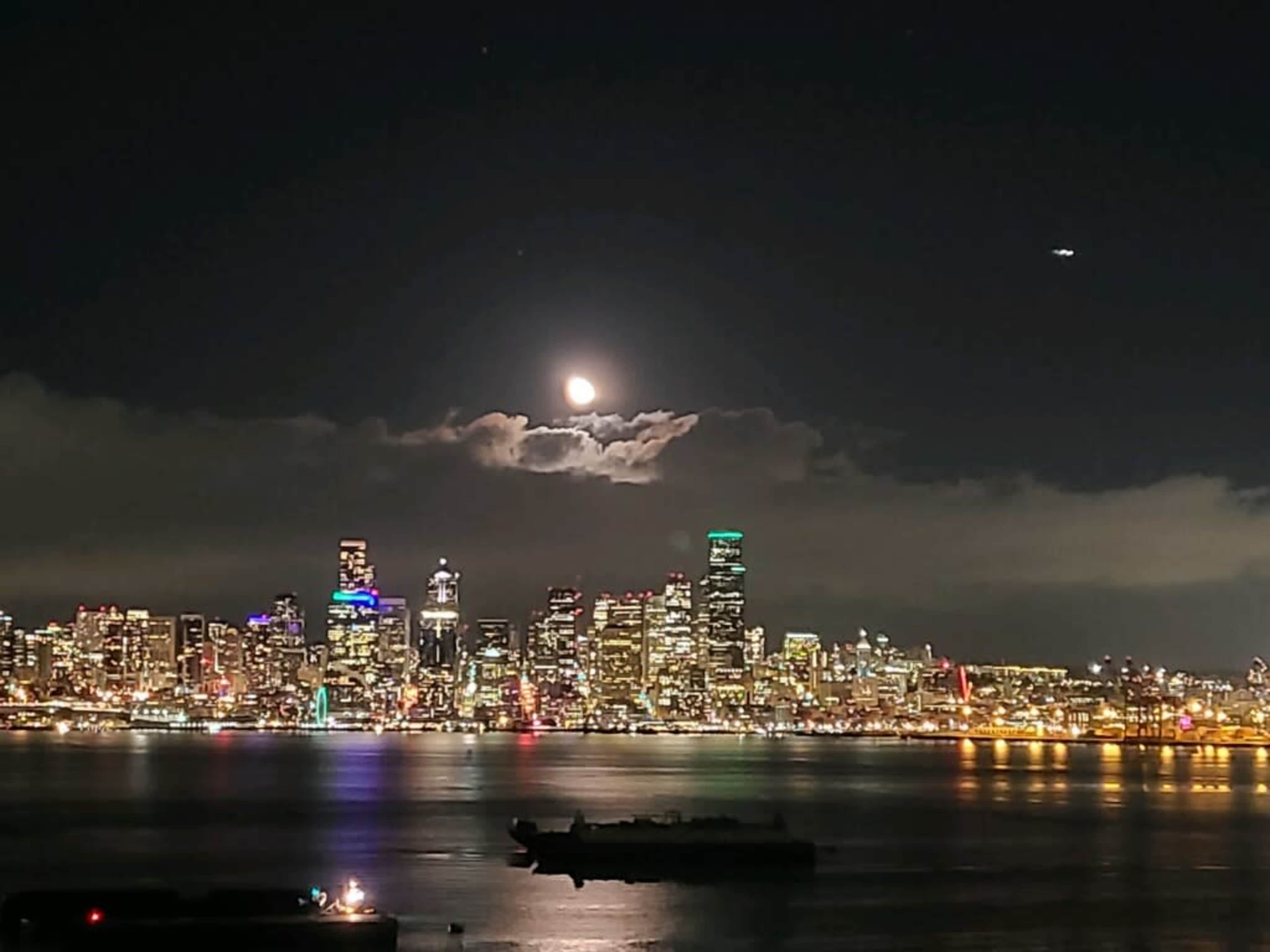 A full moon rises behind clouds over a shimmering cityscape at night, with a ship in the foreground.