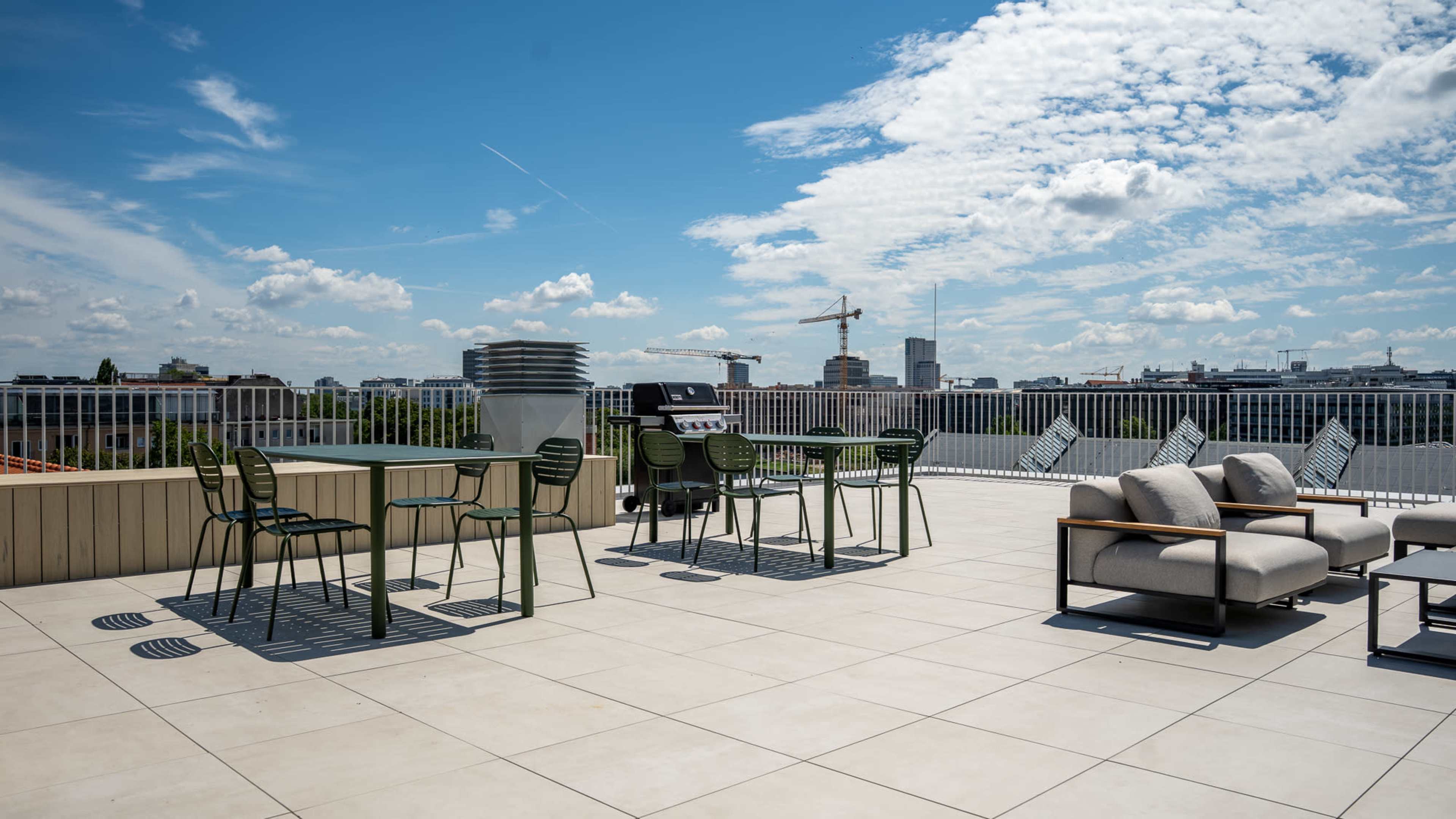 The image shows a rooftop terrace with chairs and a grill, overlooking a cityscape under a partly cloudy sky.