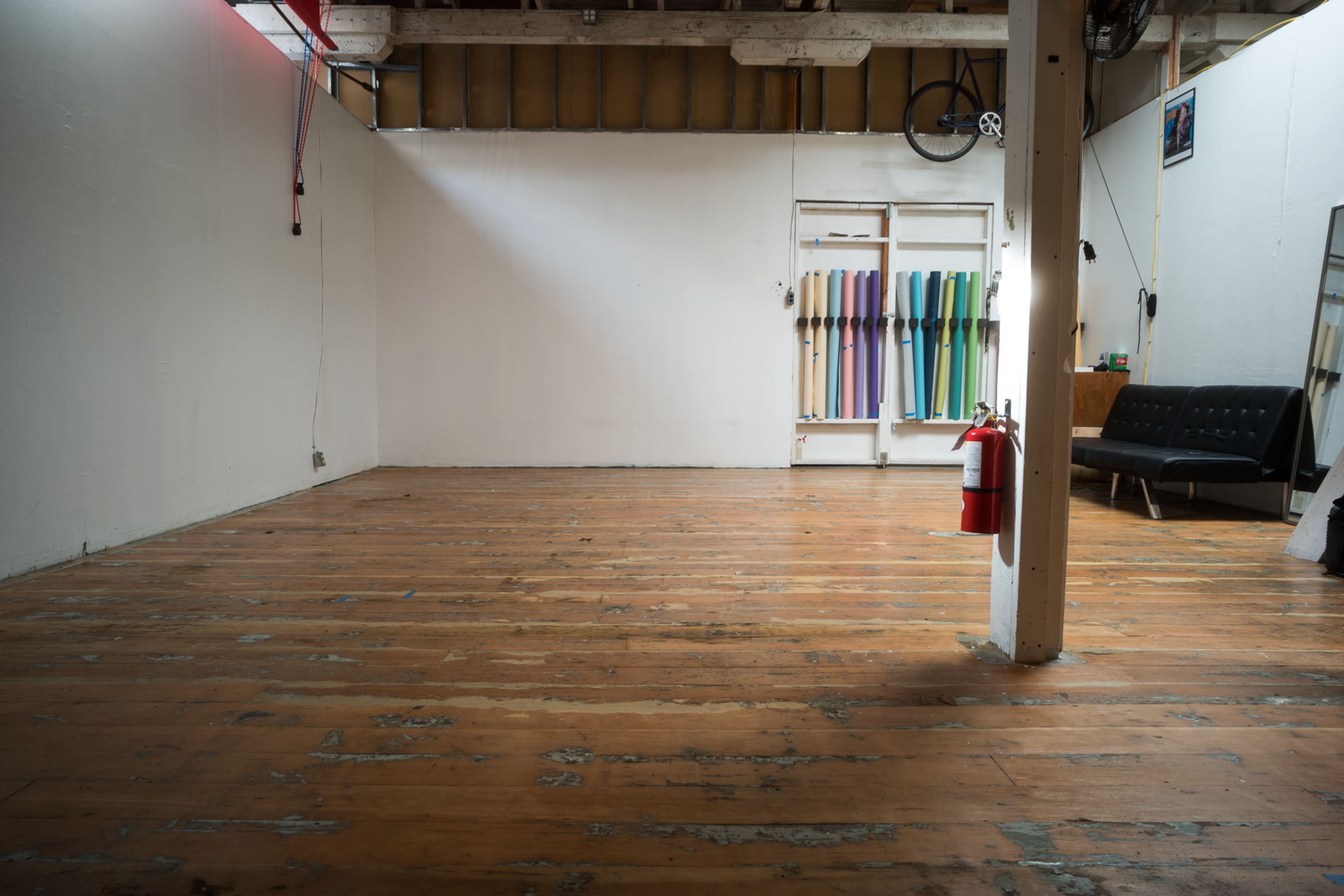An empty room features wooden floors, a white painted wall, a couch, a fire extinguisher, and colorful materials hanging on another wall.