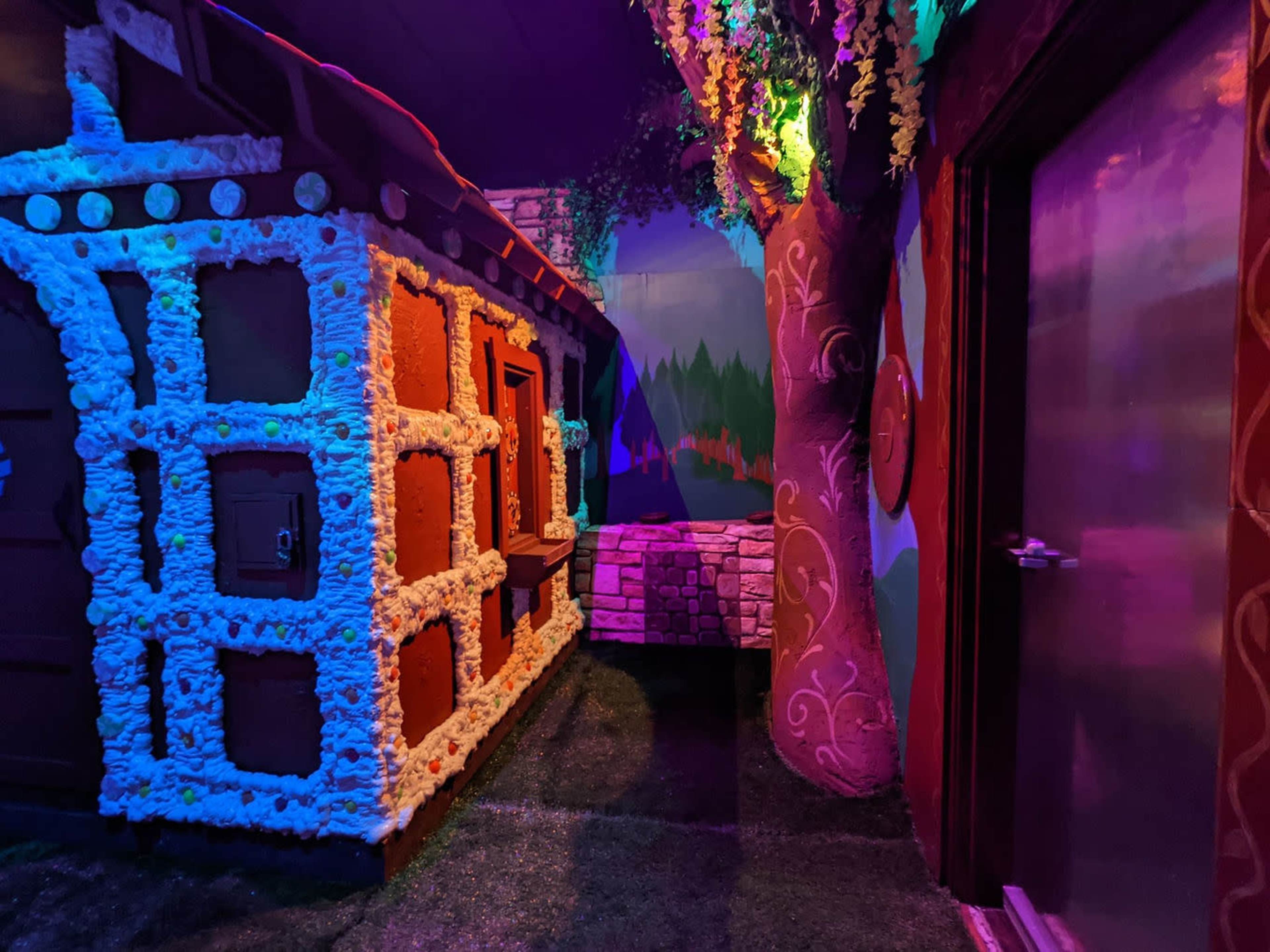 A colorful, whimsical indoor scene resembling a fairy tale environment, featuring a decorated house structure, a stone pathway, and a large tree.