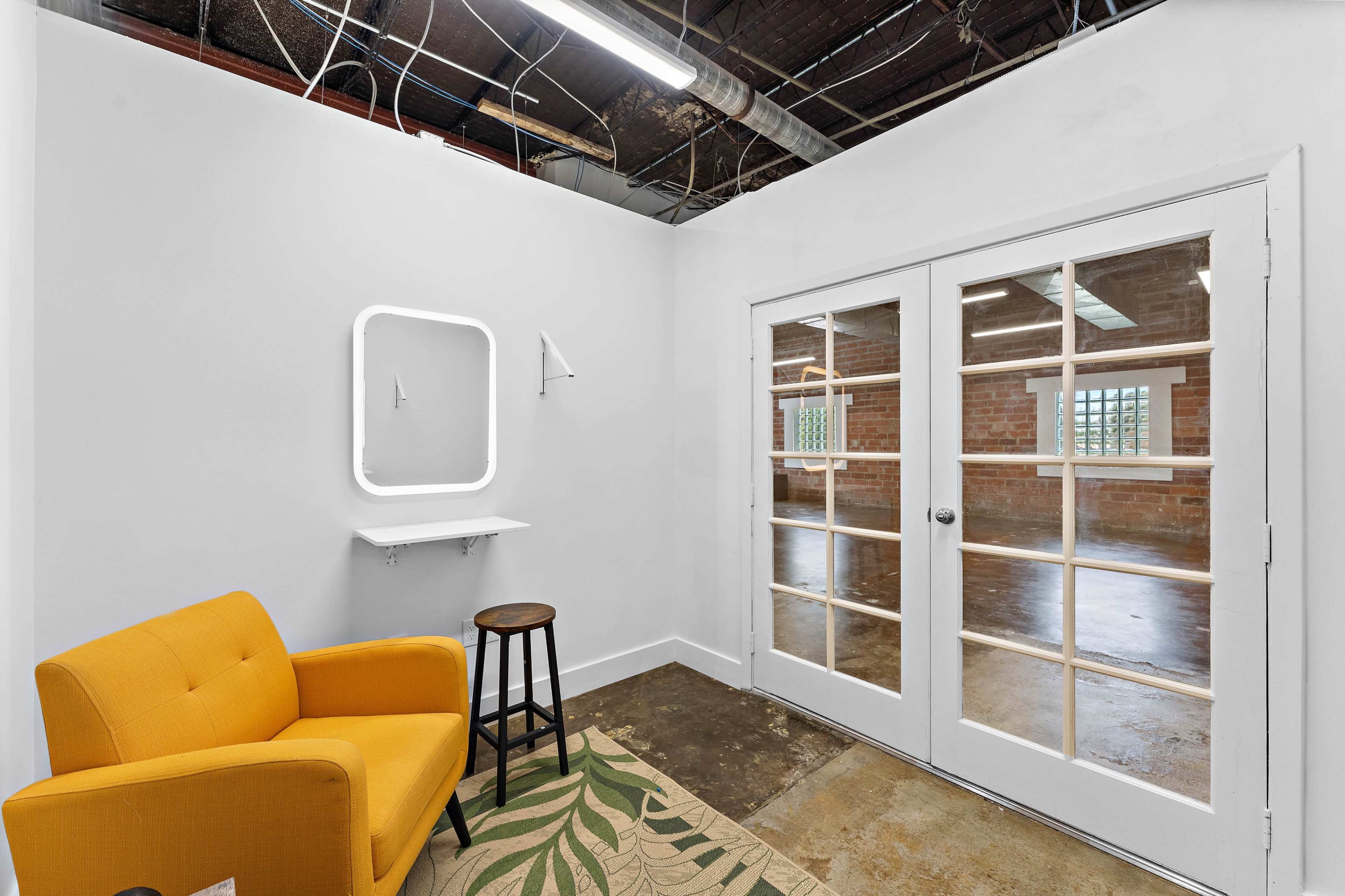 The image shows a small room featuring a yellow armchair, a wooden stool, a wall-mounted mirror, and glass-paned double doors leading to a larger, unfinished space.