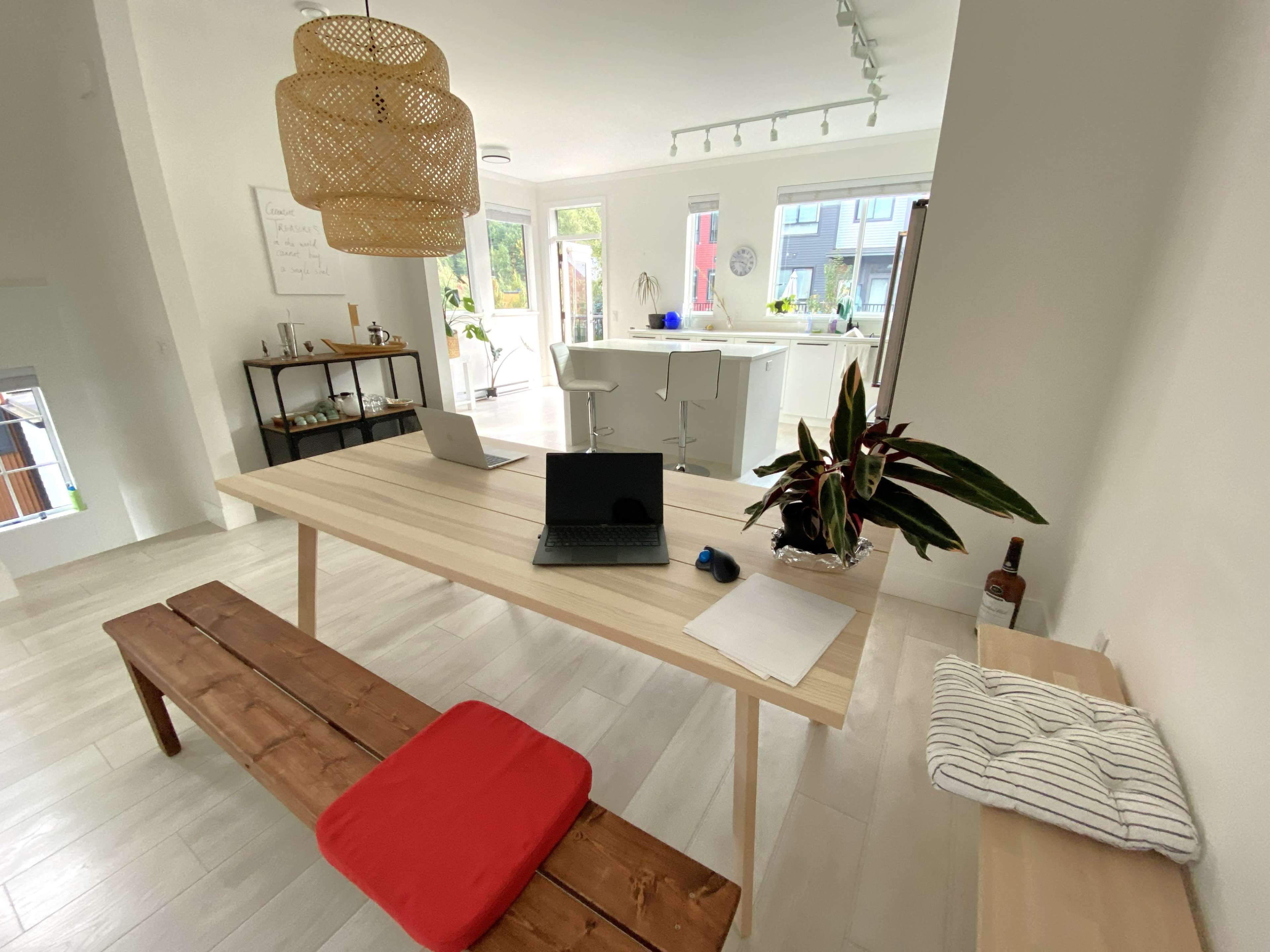 A modern interior space featuring a wooden table with a laptop, a seating bench with a red cushion, and a bright kitchen area in the background.