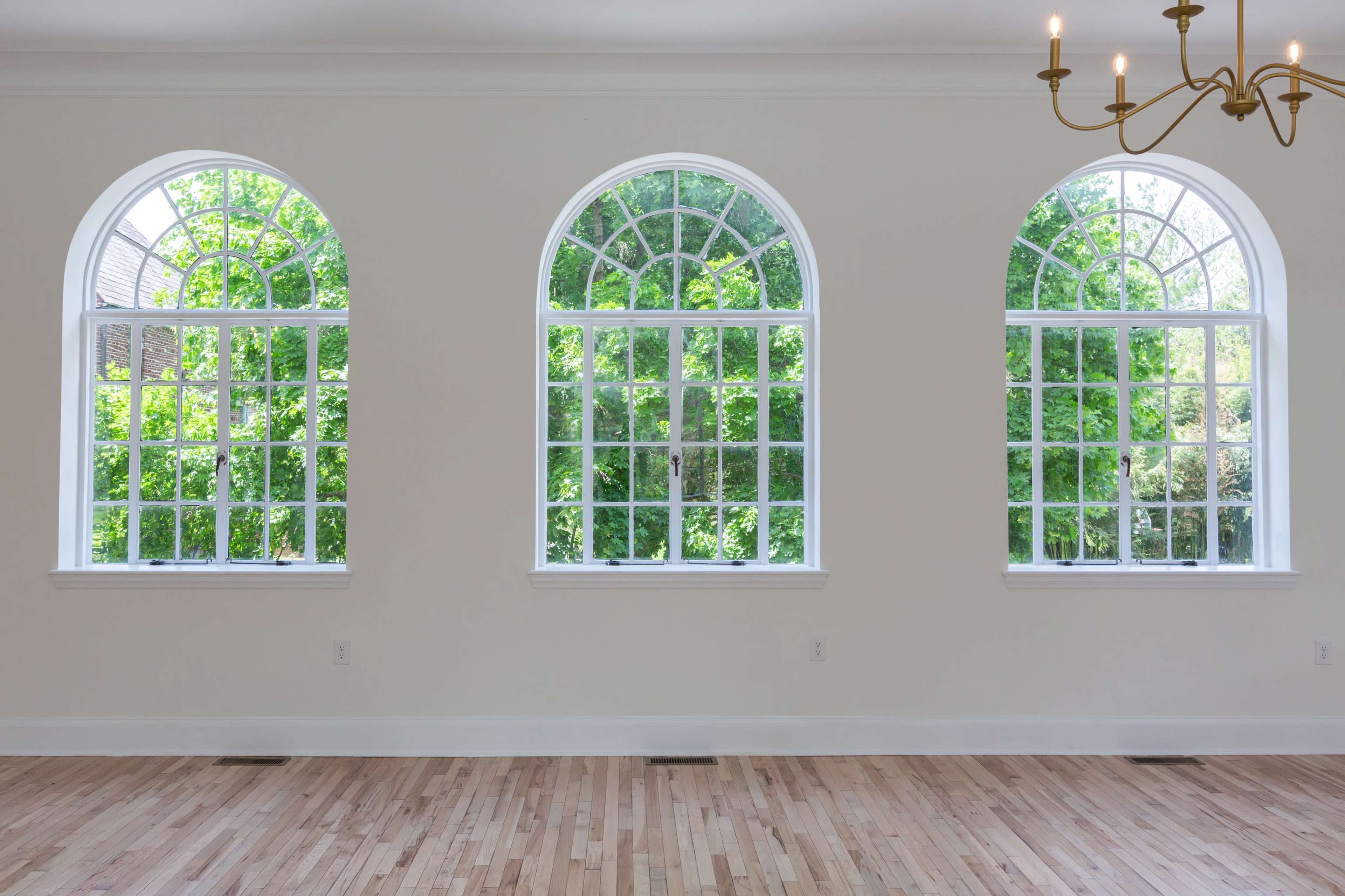 A room with three arched windows that overlook a garden, featuring wooden flooring and a chandelier overhead.