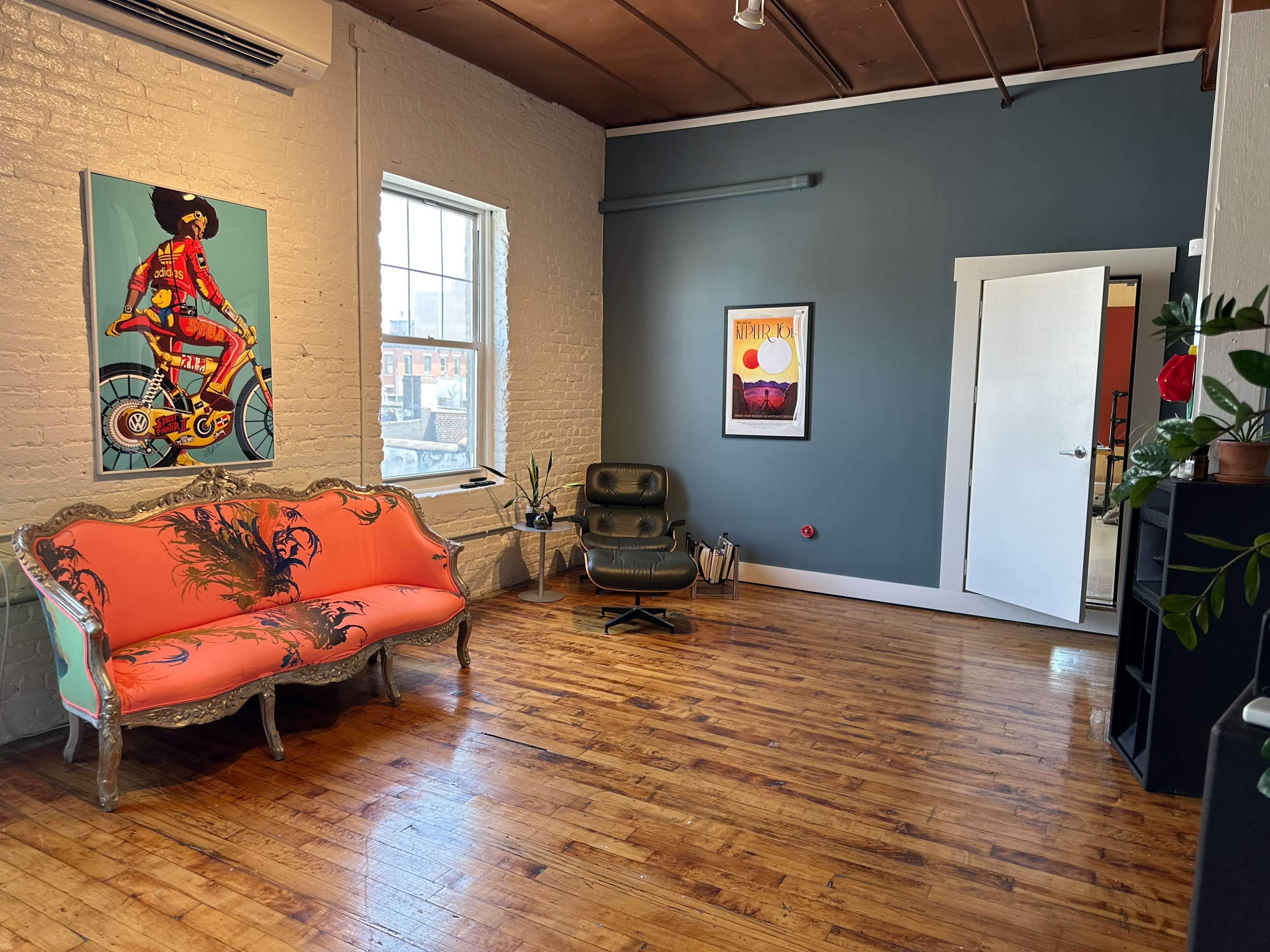 The room features a colorful sofa, a green chair, and light wooden floors, with artwork hanging on the walls and large windows providing natural light.