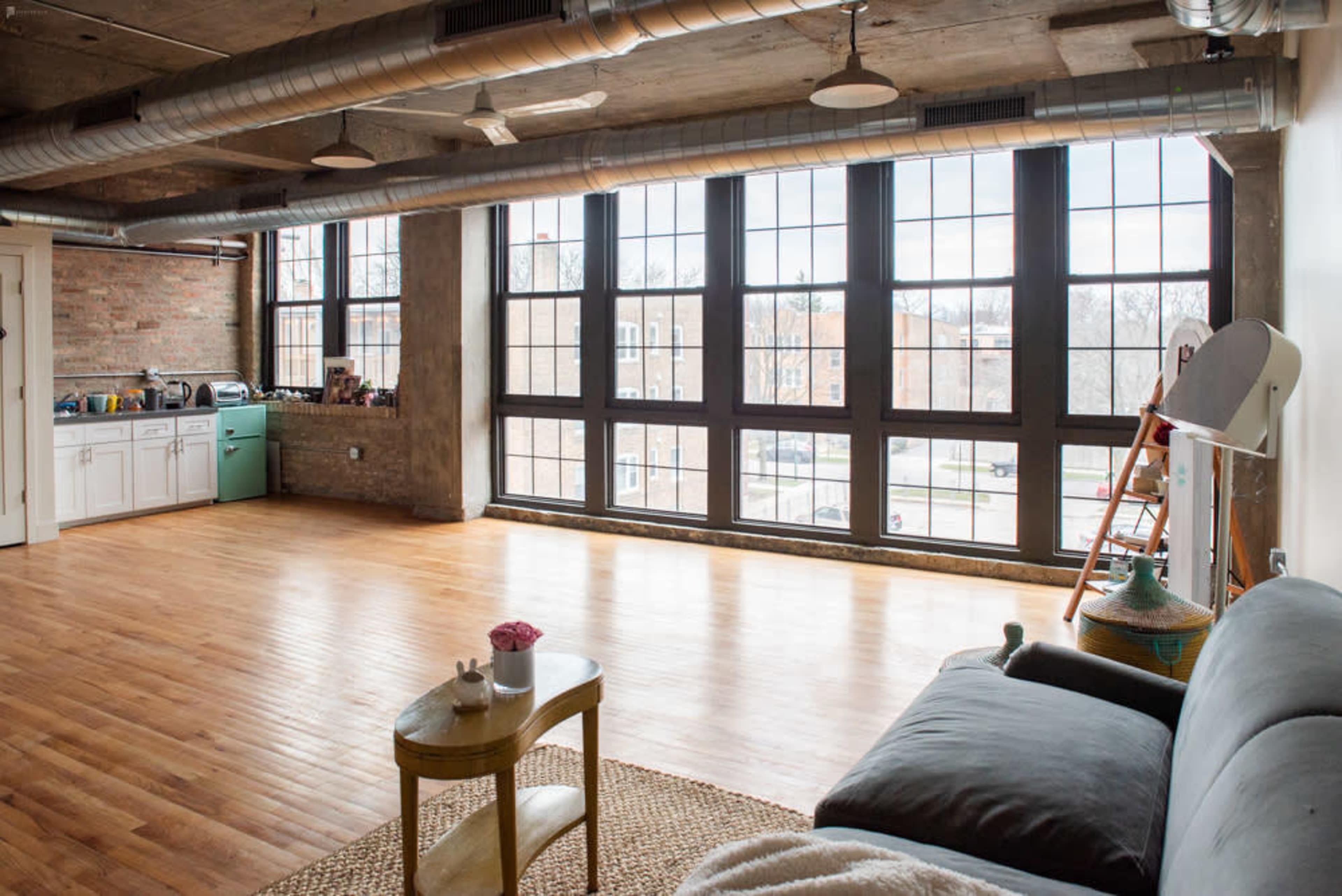 A spacious, open-concept living area features large windows with a view of the street, hardwood floors, and a kitchen area in the background.