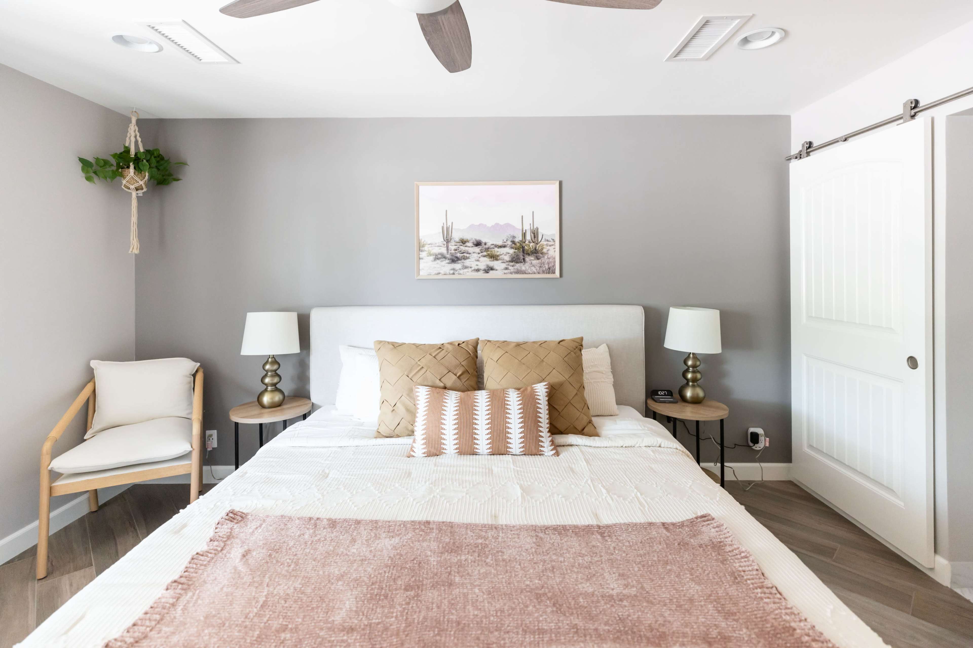 A bedroom features a bed with decorative pillows, two bedside lamps, and a framed landscape painting on the gray wall.
