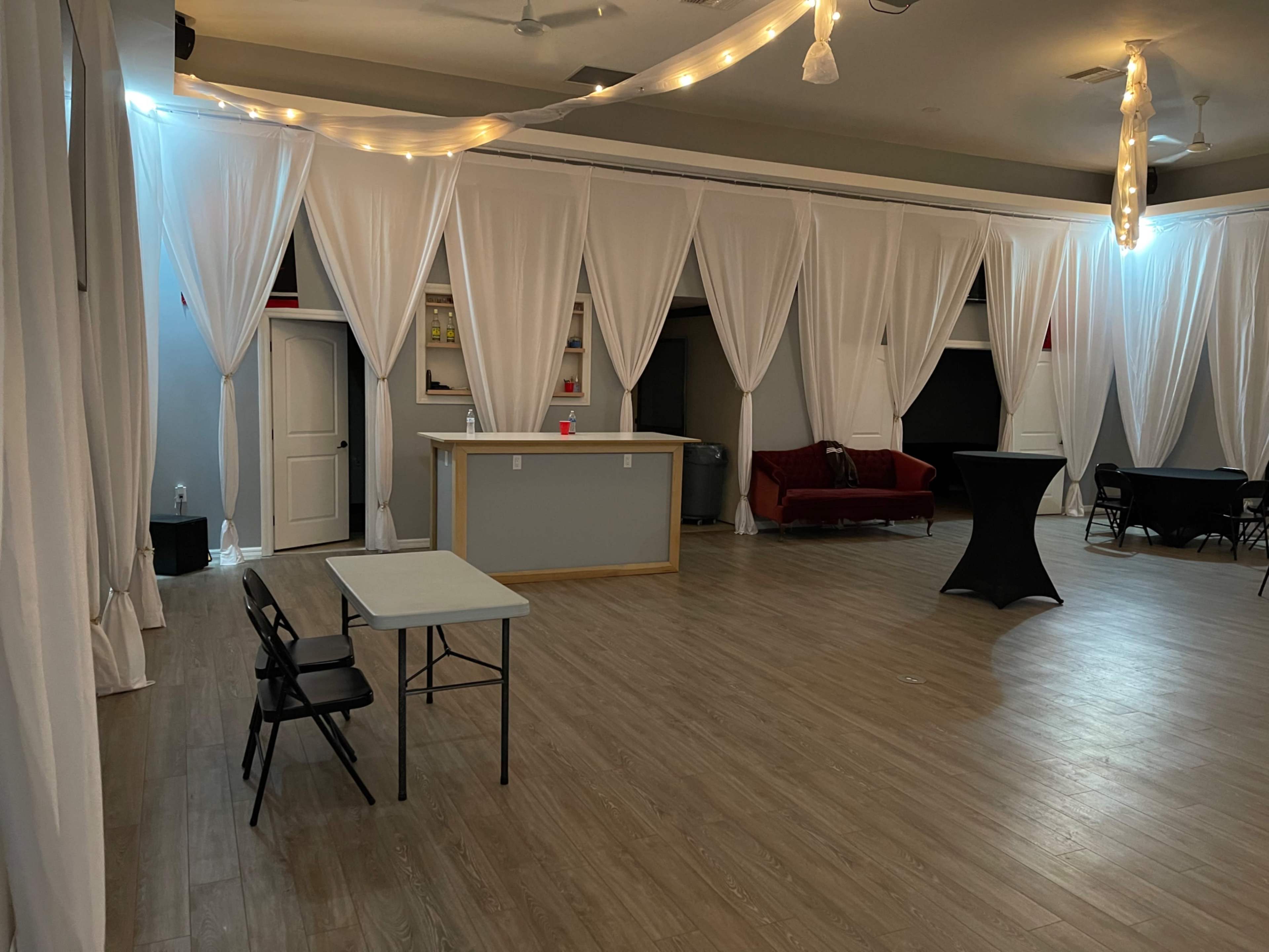 The image shows an event space with white draped curtains, a bar area, a red couch, and several tables and chairs spread throughout the room.