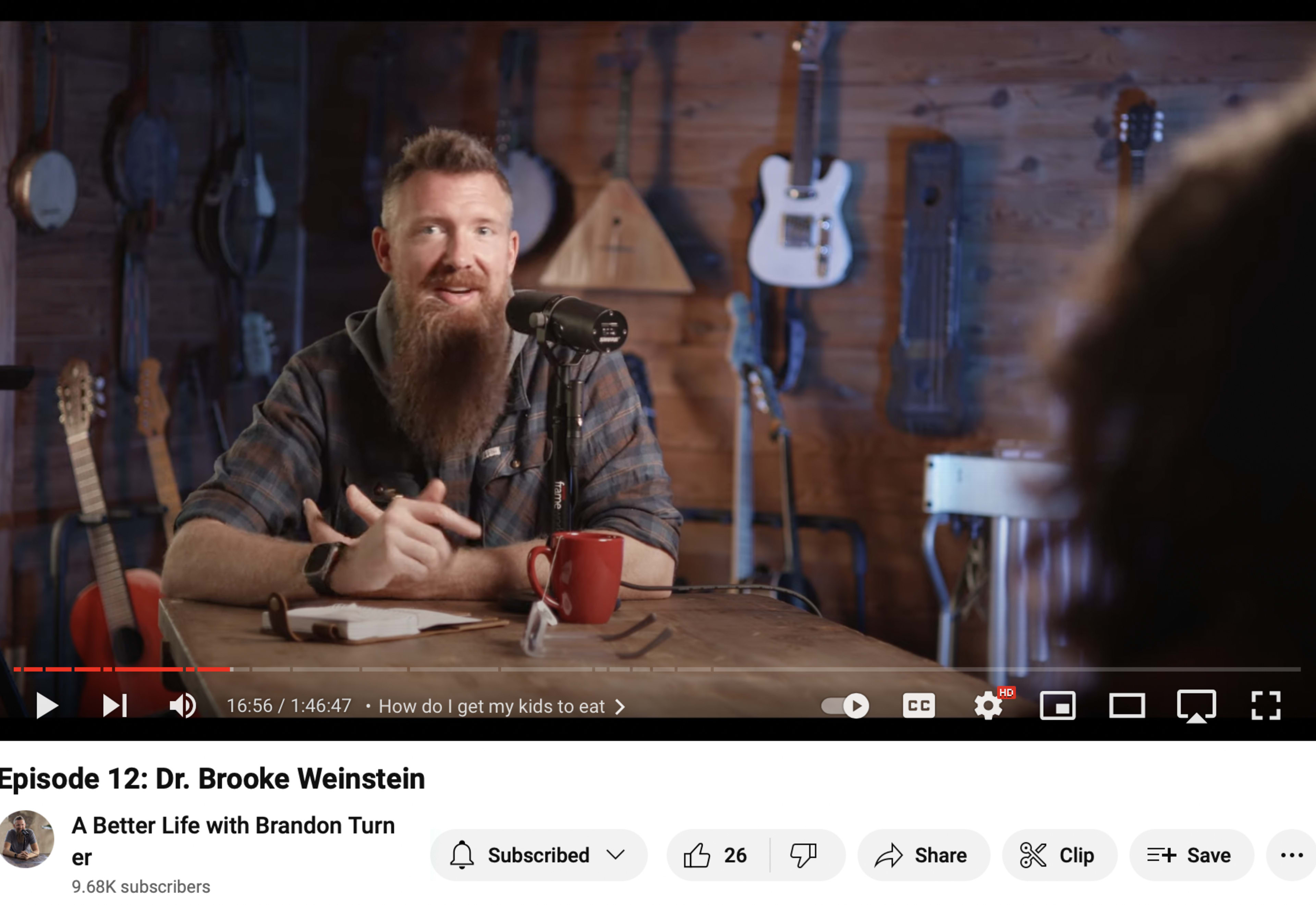 A man with a beard sits at a table in a studio decorated with various guitars, speaking into a microphone.