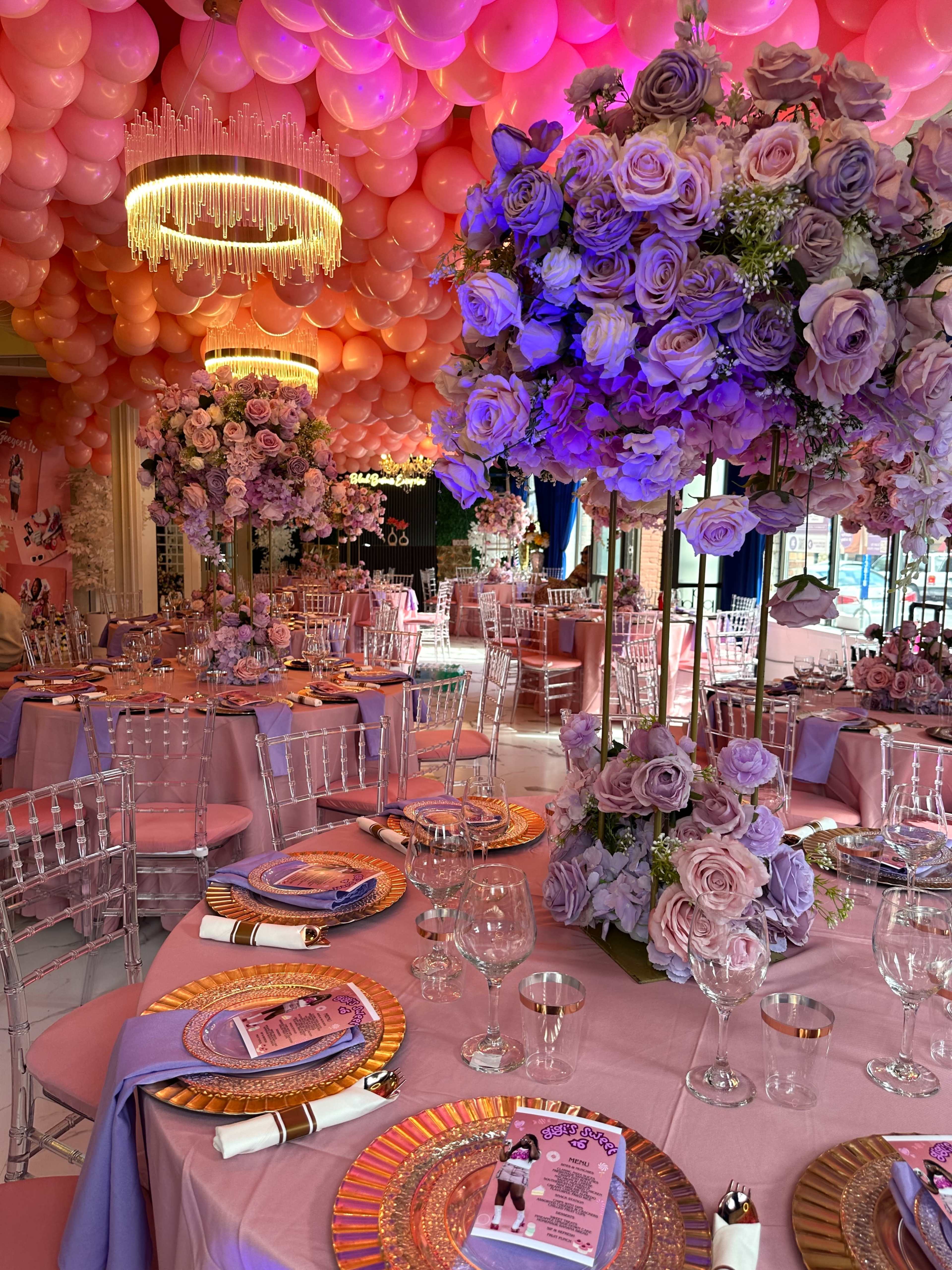 The scene features an elegant event space adorned with large floral centerpieces, pink and purple balloons overhead, and beautifully set tables with vibrant tableware.
