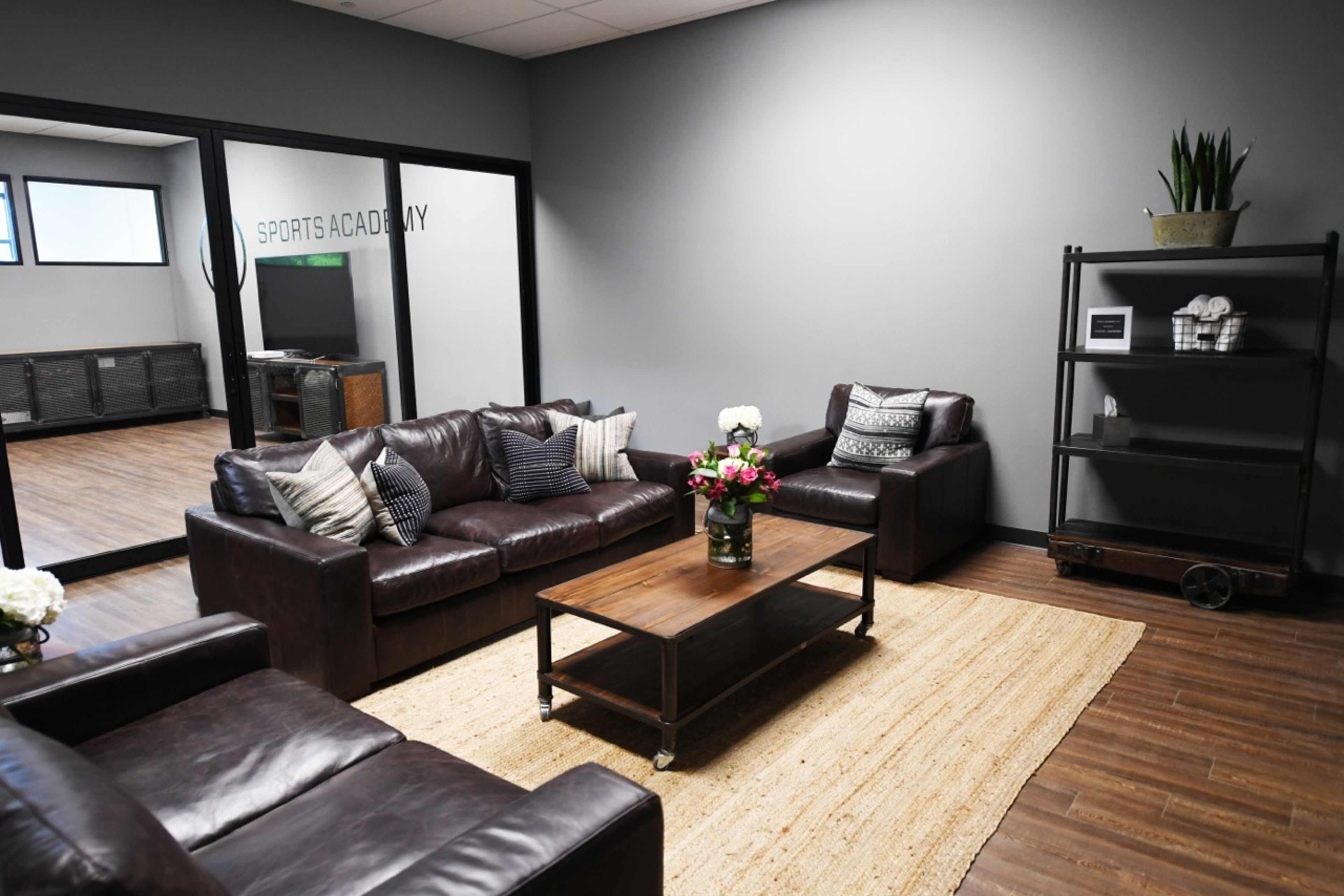 A waiting room with brown leather sofas, a wooden coffee table with a floral arrangement, and a black shelving unit against a gray wall.