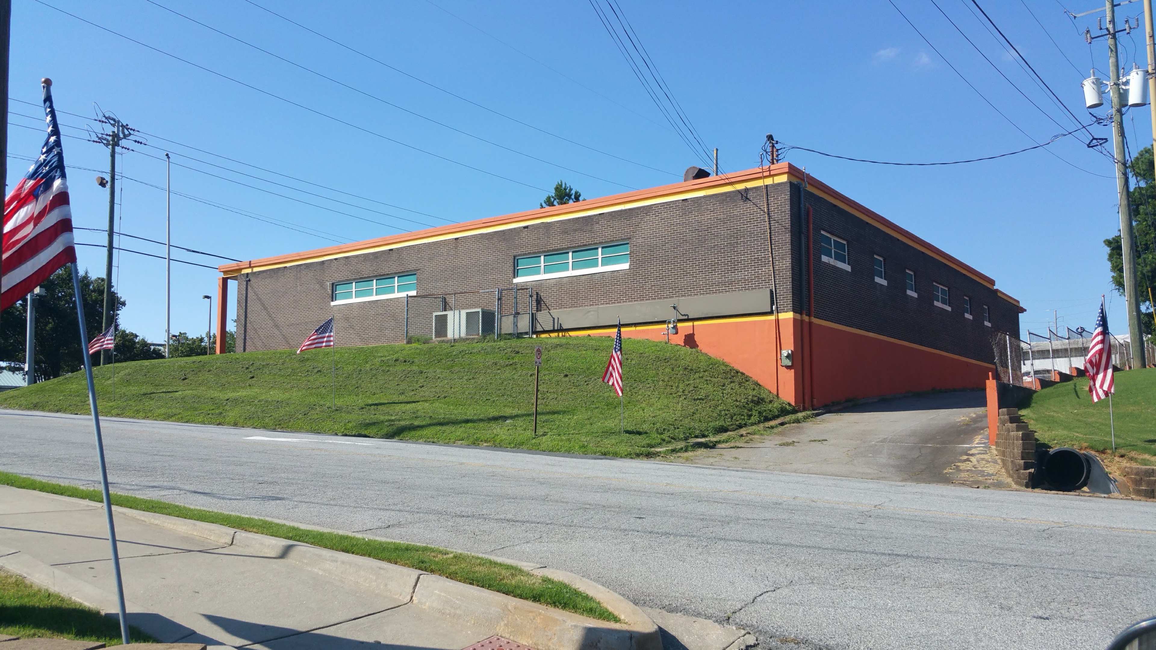 The image shows a brick building with an orange trim located on a grassy slope, flanked by small American flags along the roadside.