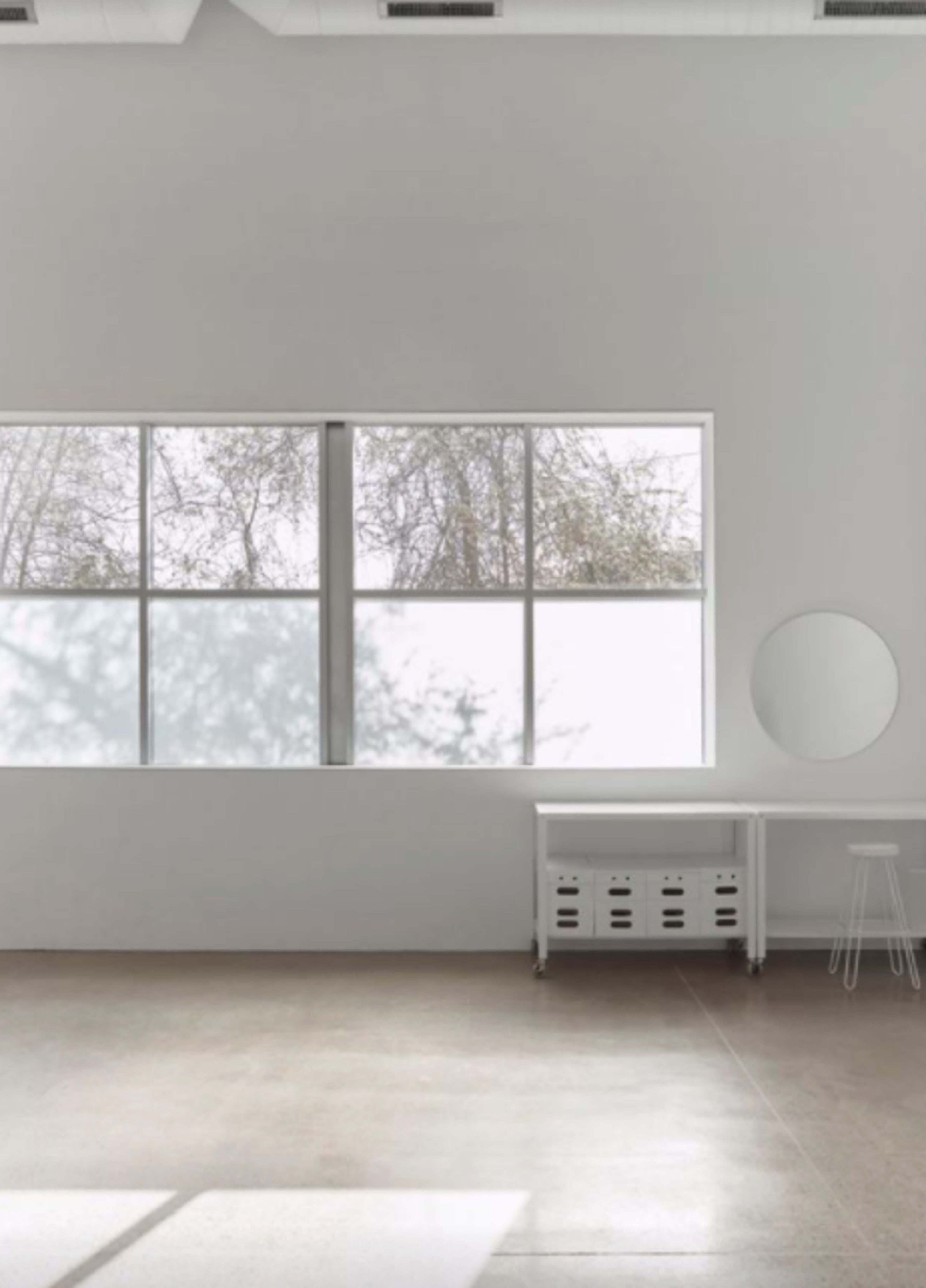 A minimalist room features a large window with trees visible outside, a circular mirror on the wall, a table with storage boxes, and a stool.