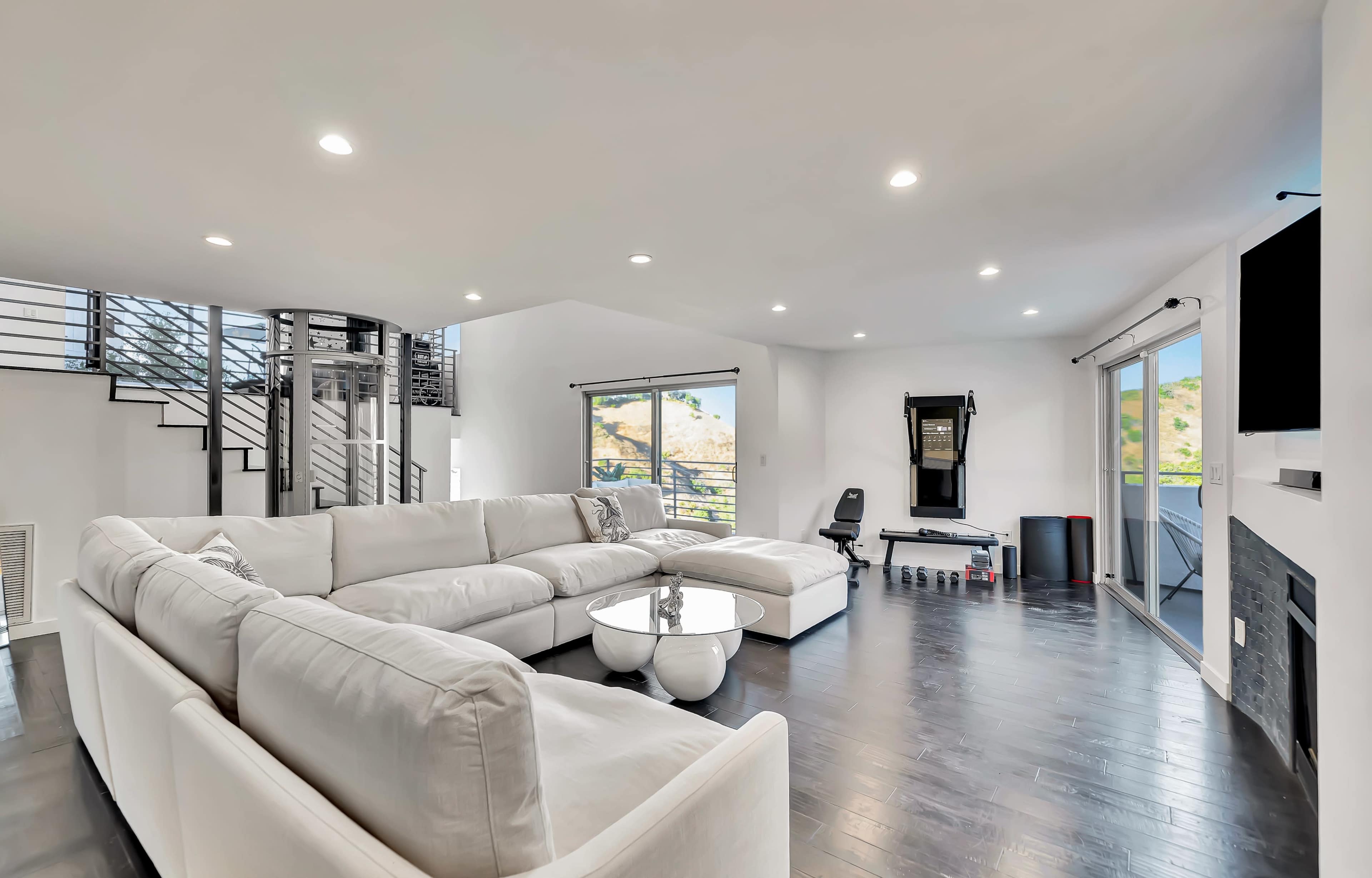 The image shows a modern, spacious living room with a large sectional sofa, large windows, and a staircase in the background.