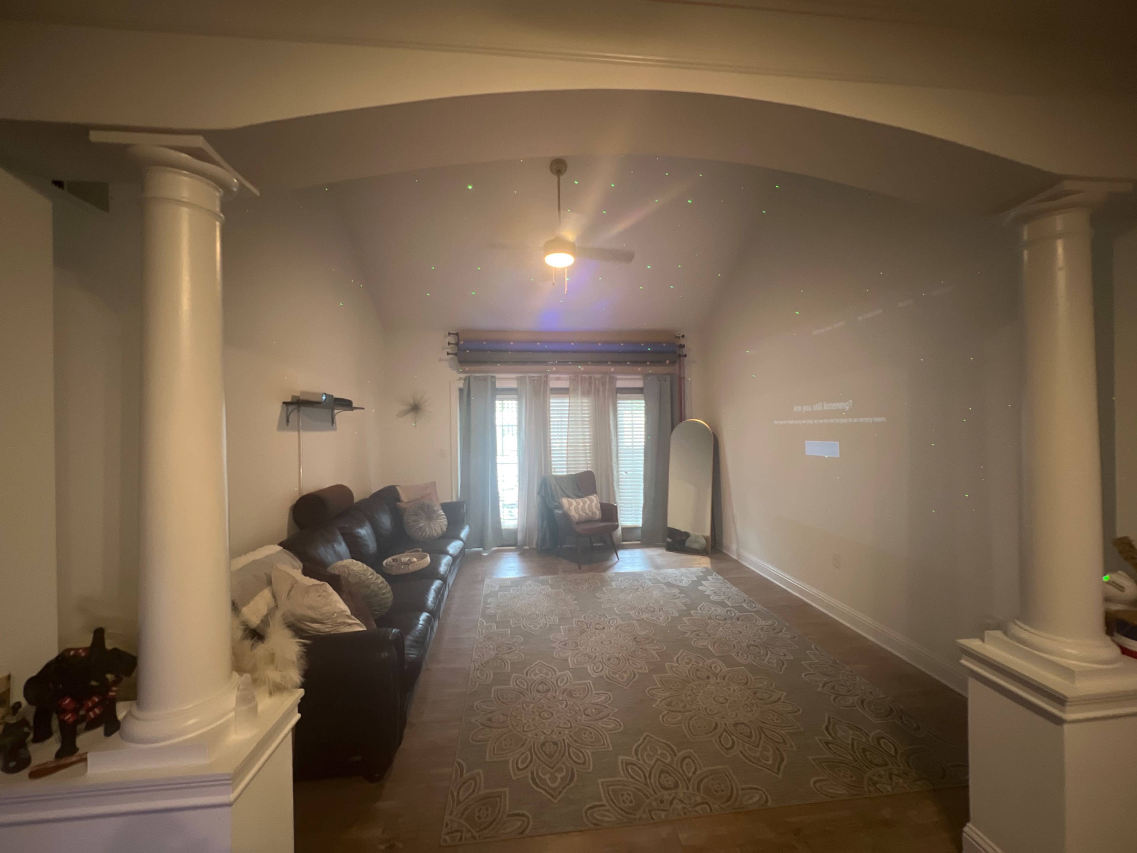 The living room features a leather sofa, a mirror, and decorative lighting projected on the wall, with large windows draped in sheer curtains.