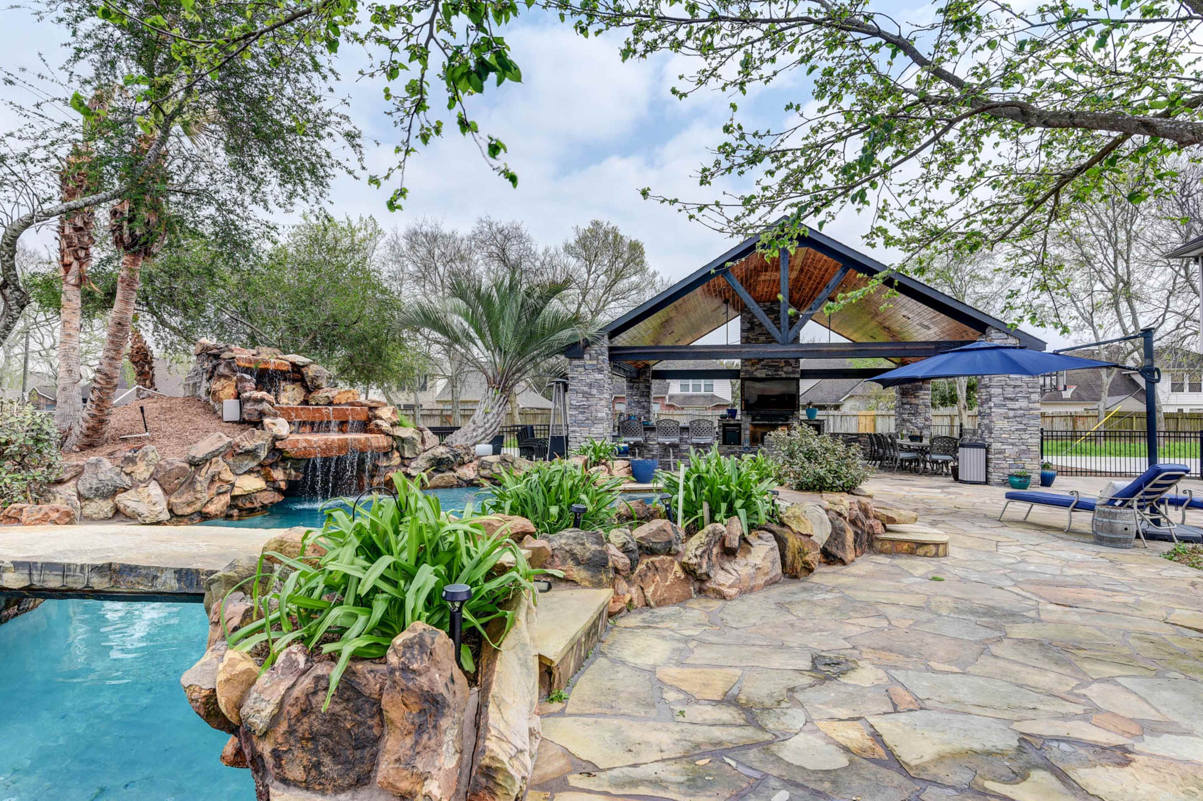 A landscaped backyard with a stone pool, a waterfall, and a covered outdoor kitchen area surrounded by greenery.