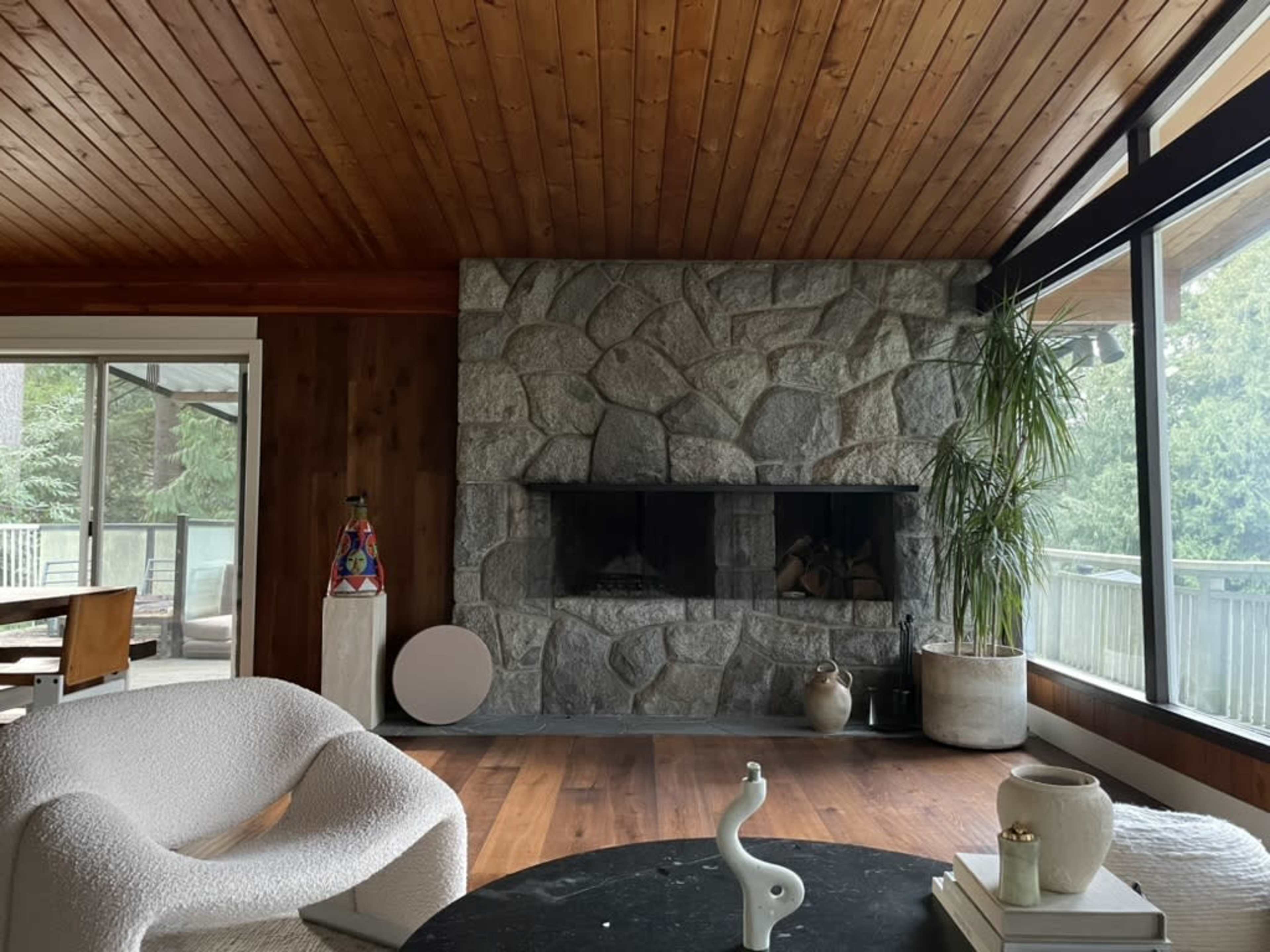The image shows a cozy living room featuring a stone fireplace, wooden ceiling, and large windows overlooking greenery.