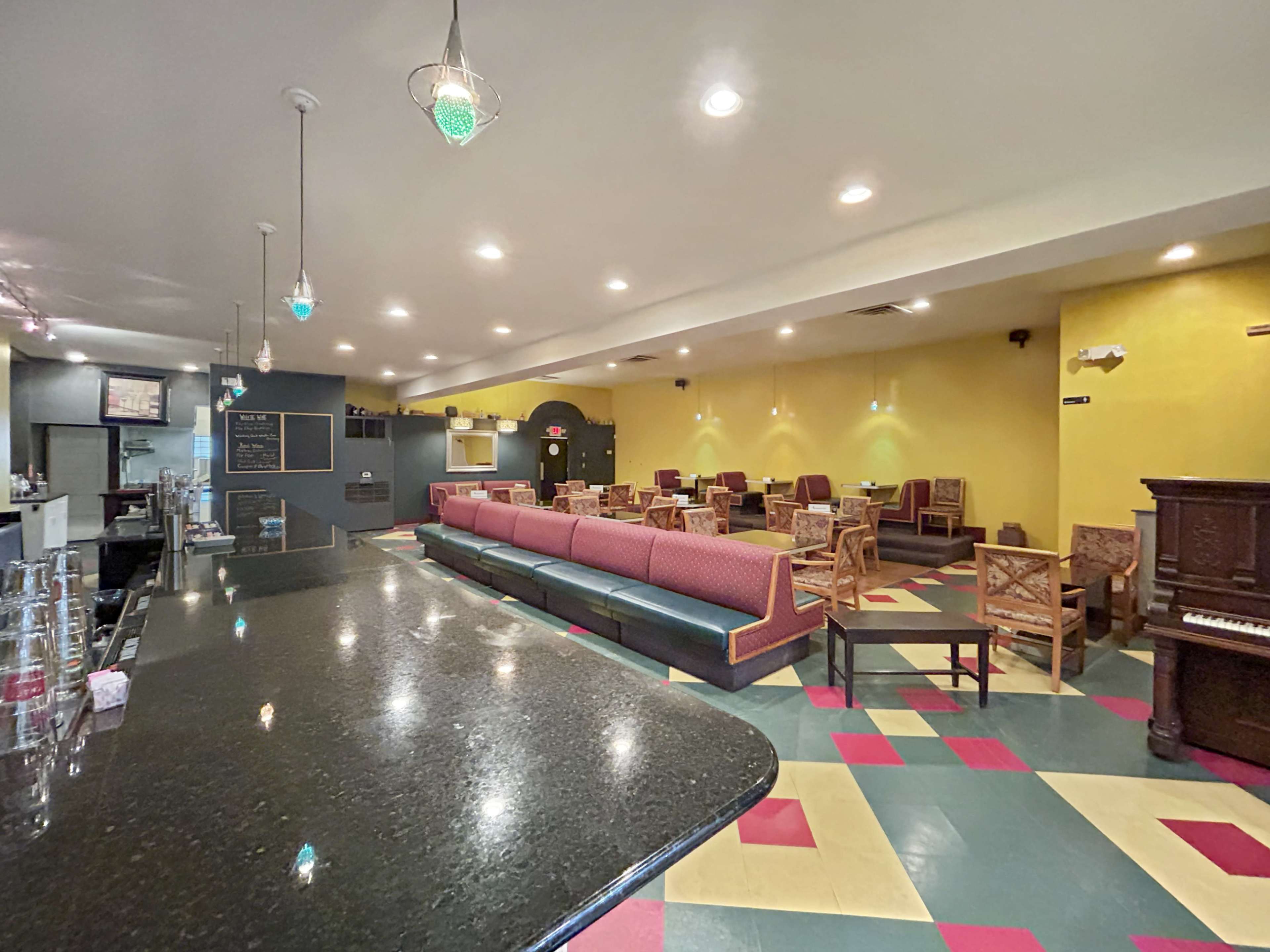 A spacious café interior featuring a bar counter, rows of upholstered seating, scattered wooden chairs, and a piano against a brightly colored wall.
