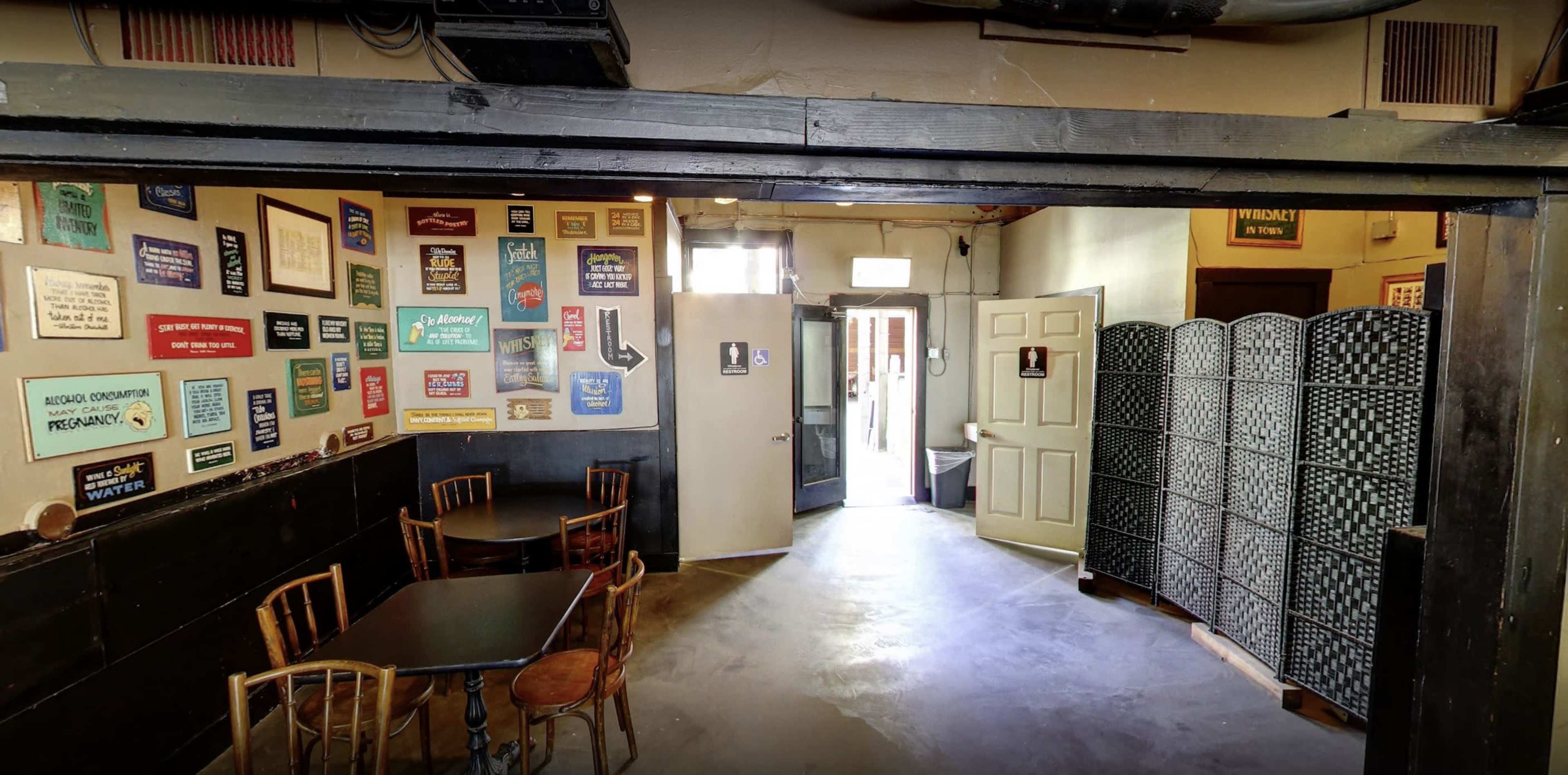 A rustic interior of a room with a series of colorful signs on the wall, tables and chairs arranged, and a door leading outside.