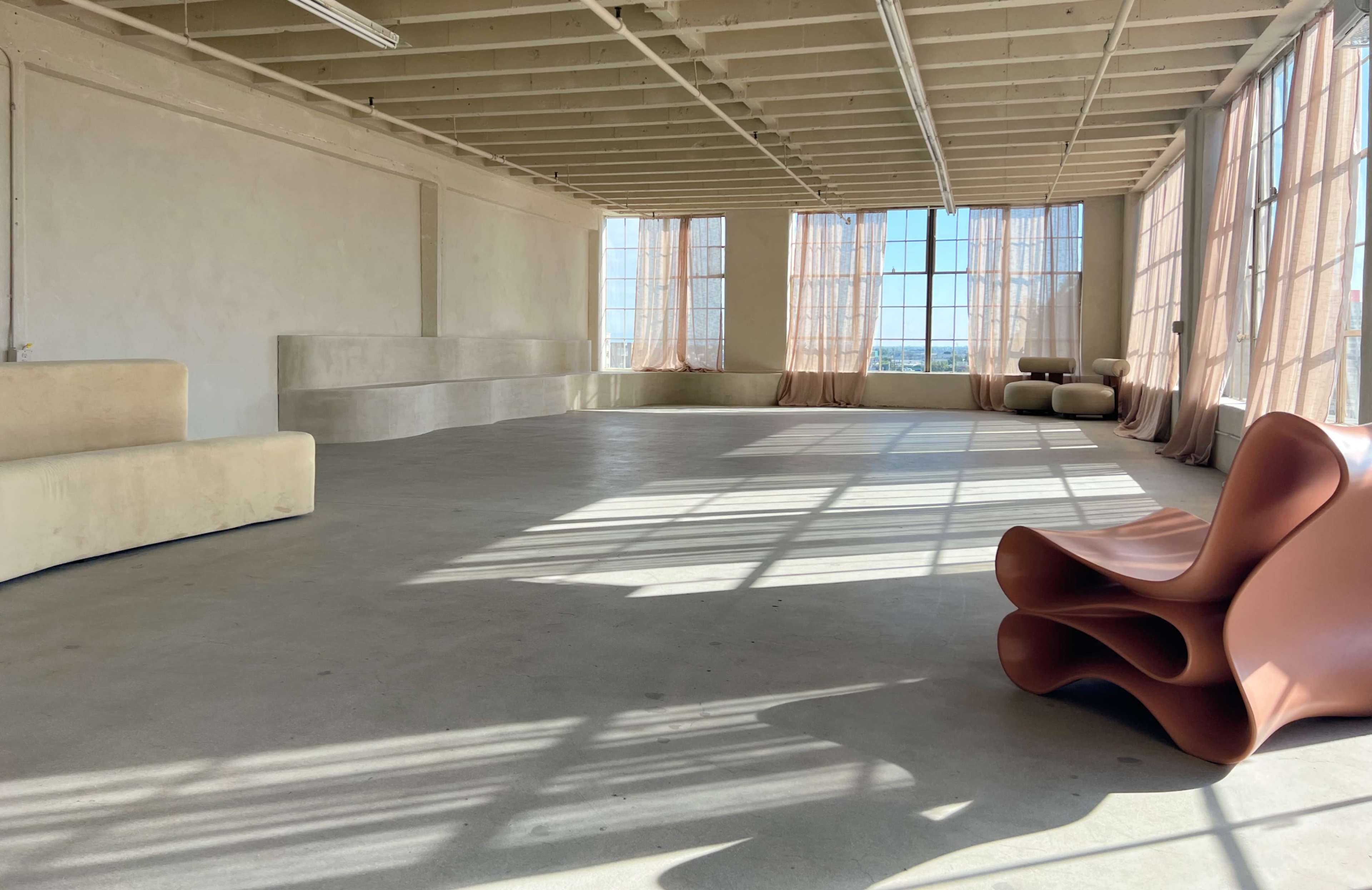 The image shows a spacious, sunlit room with large windows, minimalistic furniture, and soft, flowing curtains.