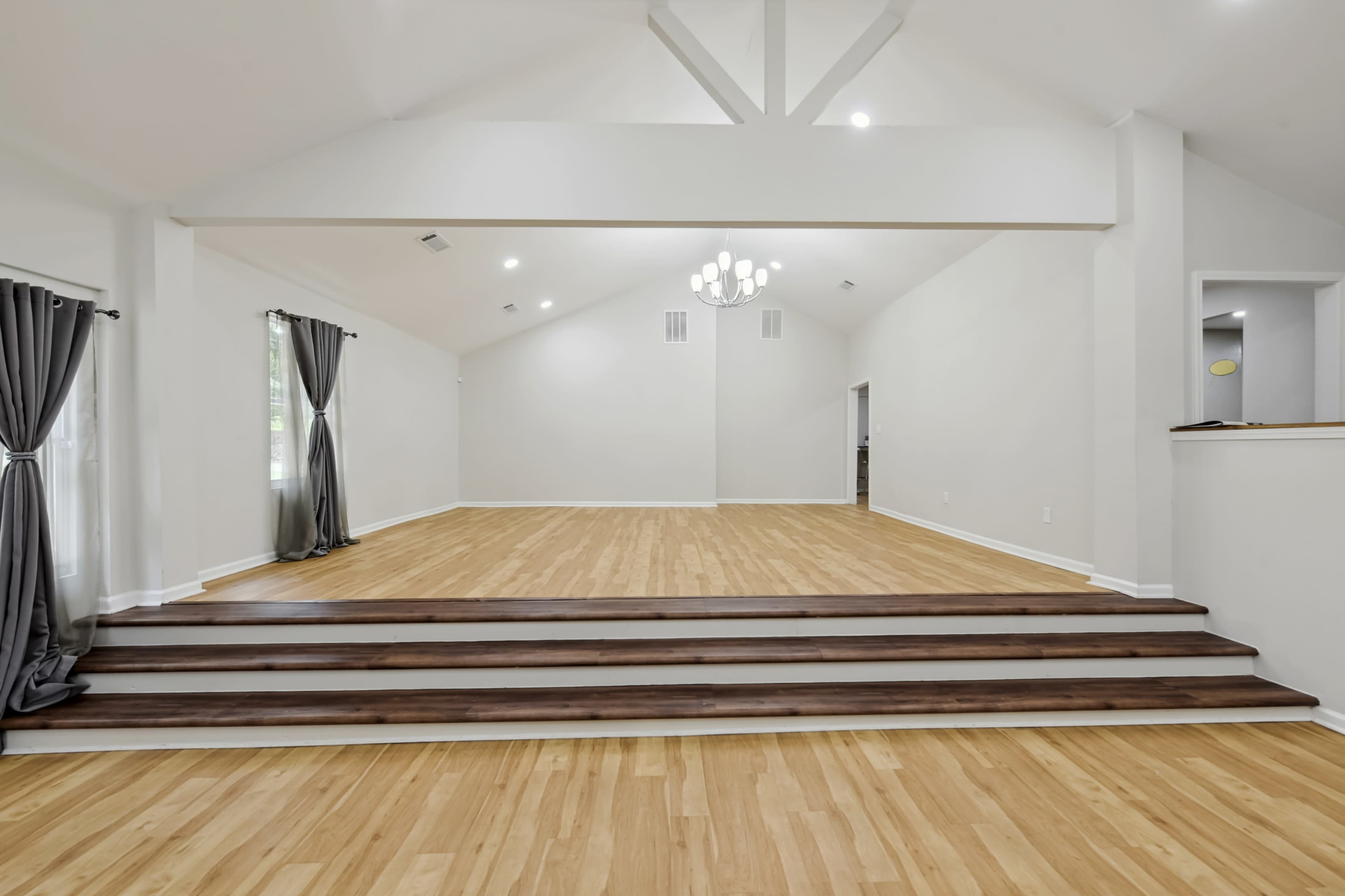 The image shows a spacious room with a high ceiling, light wood flooring, and a staircase leading up to a raised area.