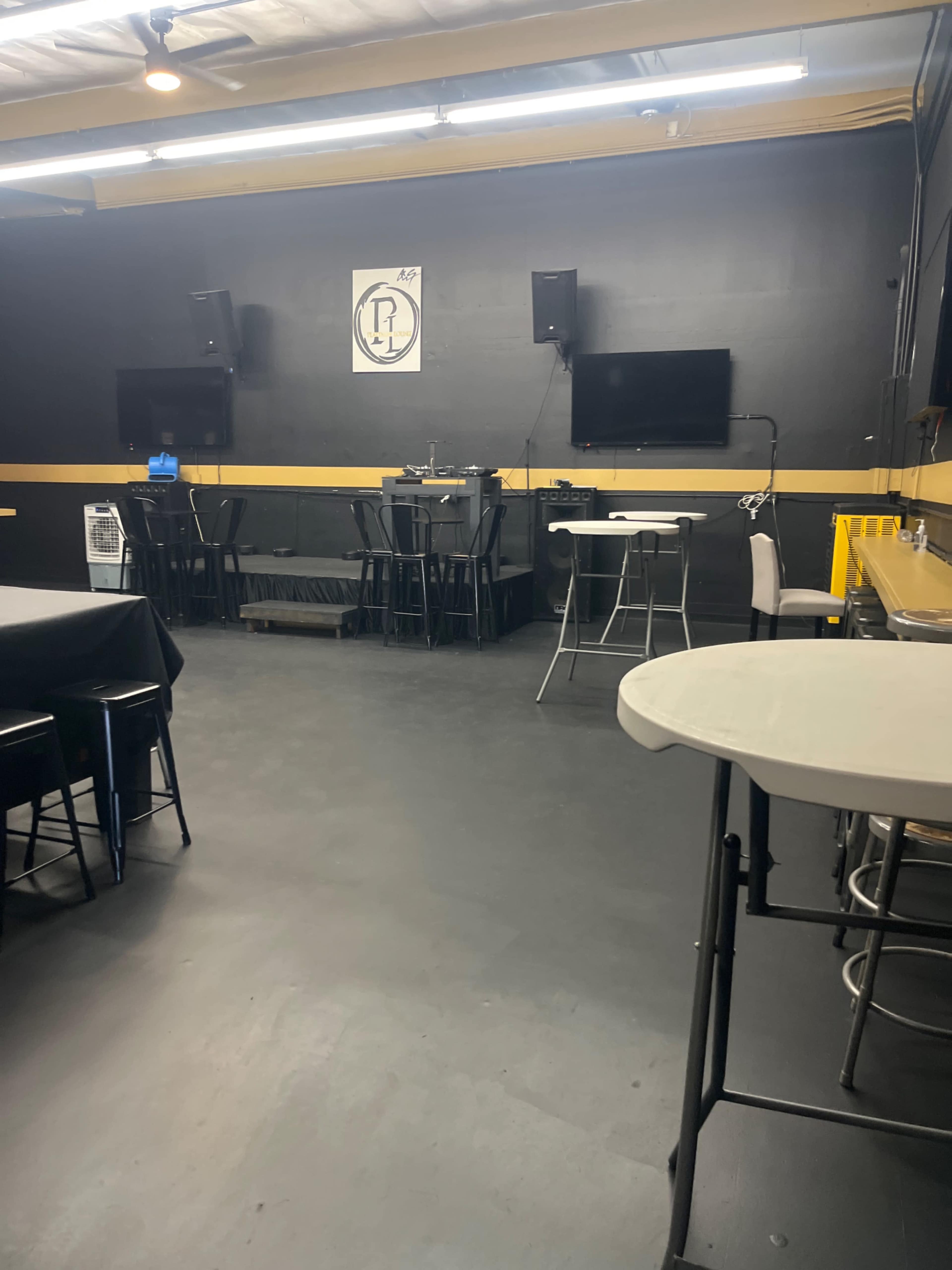 The image shows an interior space with black walls, several tables and chairs, and two wall-mounted screens.