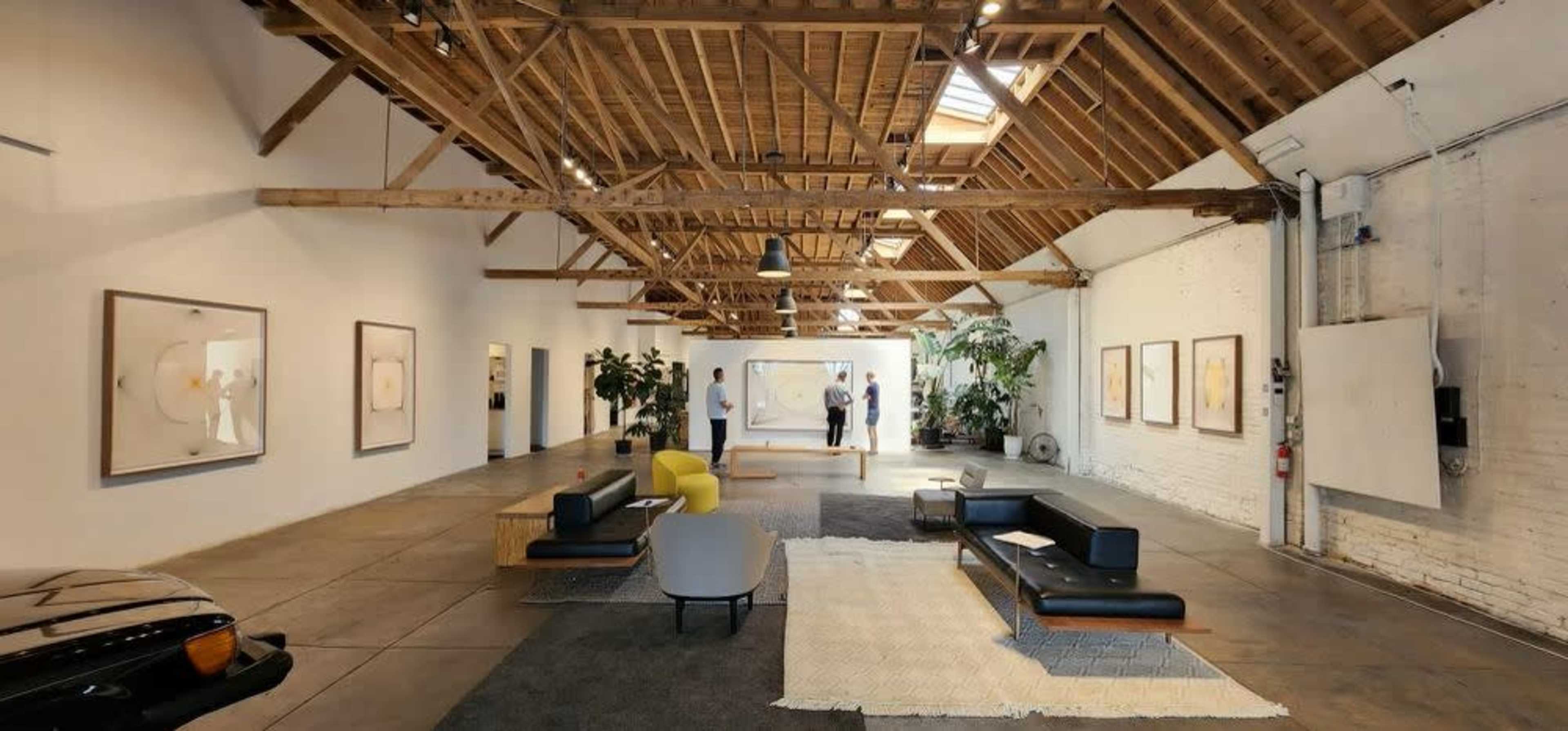 A spacious art gallery with a wooden ceiling, featuring various artworks on the walls and modern furniture arranged on the floor.