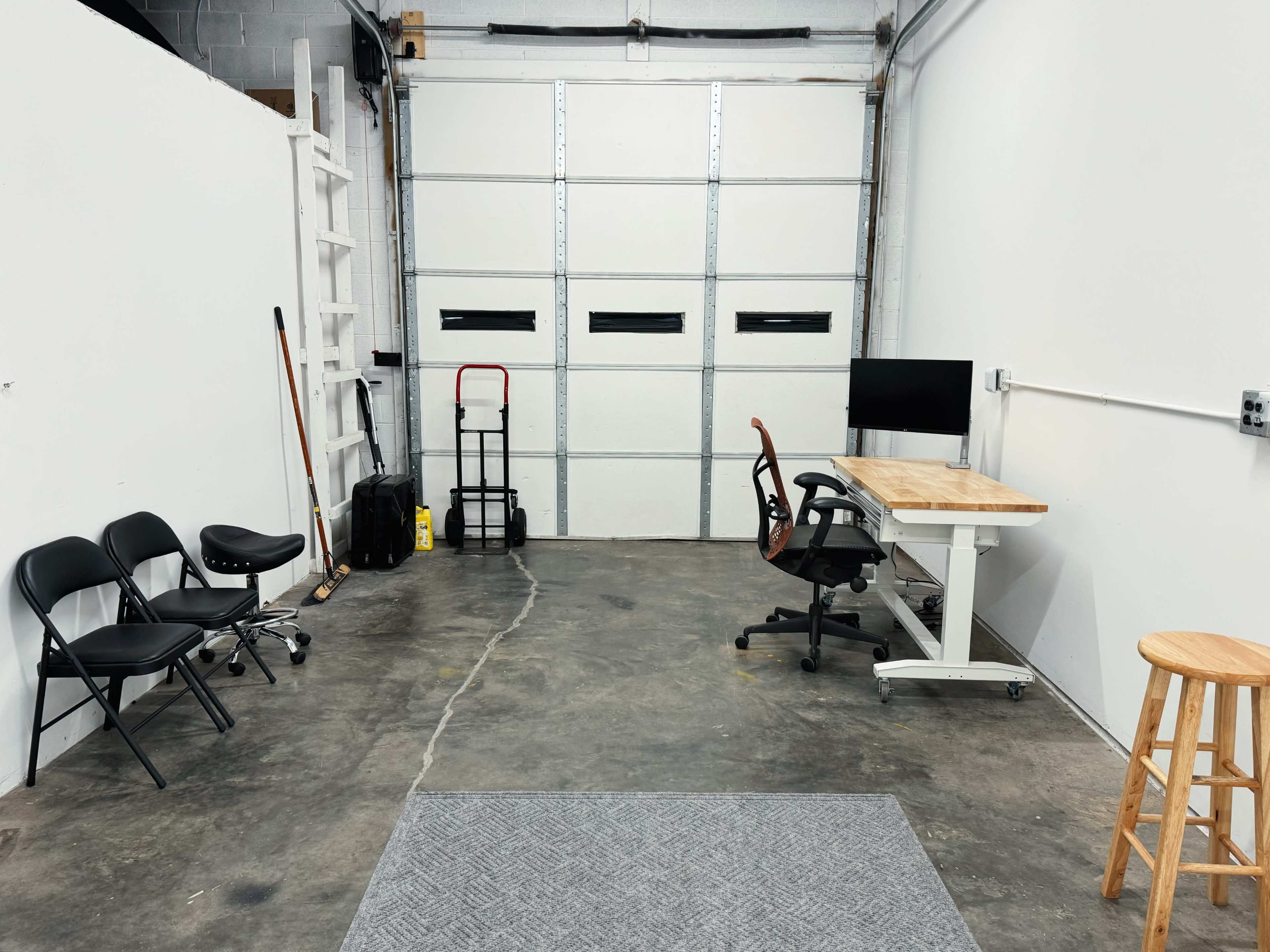 A sparse workspace with two chairs and a stool positioned next to a desk with a computer, opposite a garage door and additional empty space.