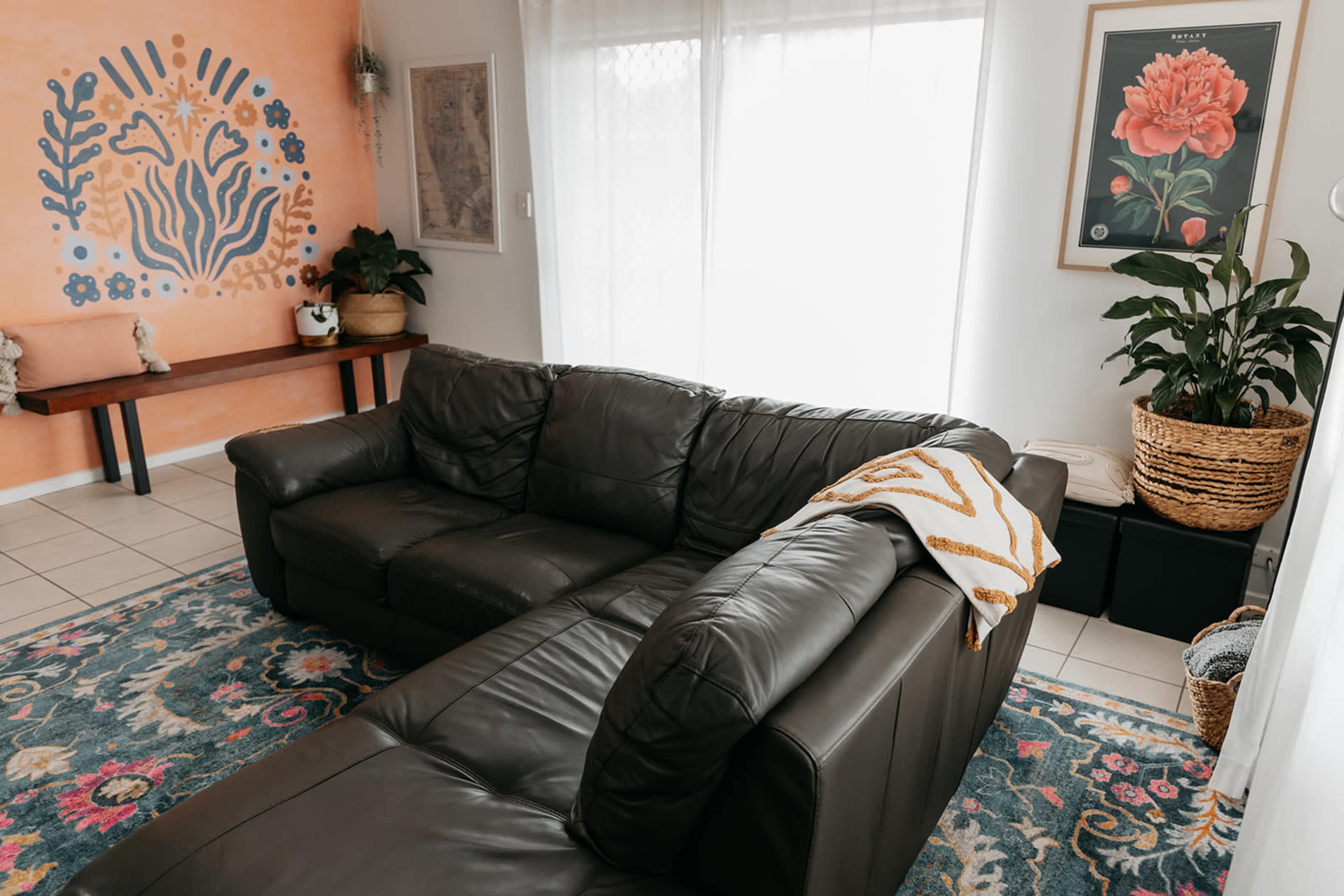 A sunlit living room featuring a dark leather sectional sofa, a decorative wall with a floral mural, and a vibrant area rug.