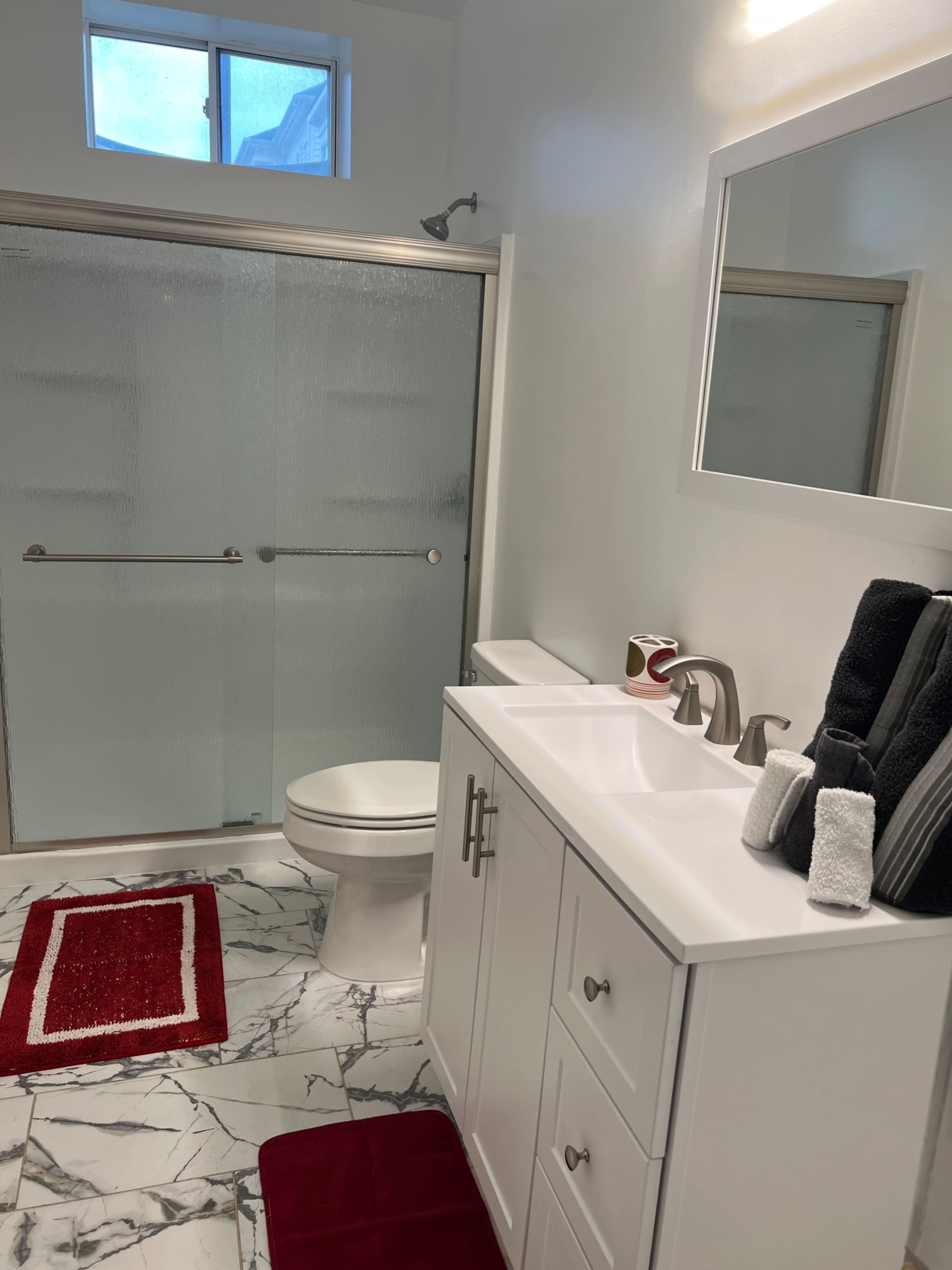 The image shows a modern bathroom featuring a glass shower enclosure, a white vanity with a sink, and marble-patterned flooring, complemented by red bath mats and towels.
