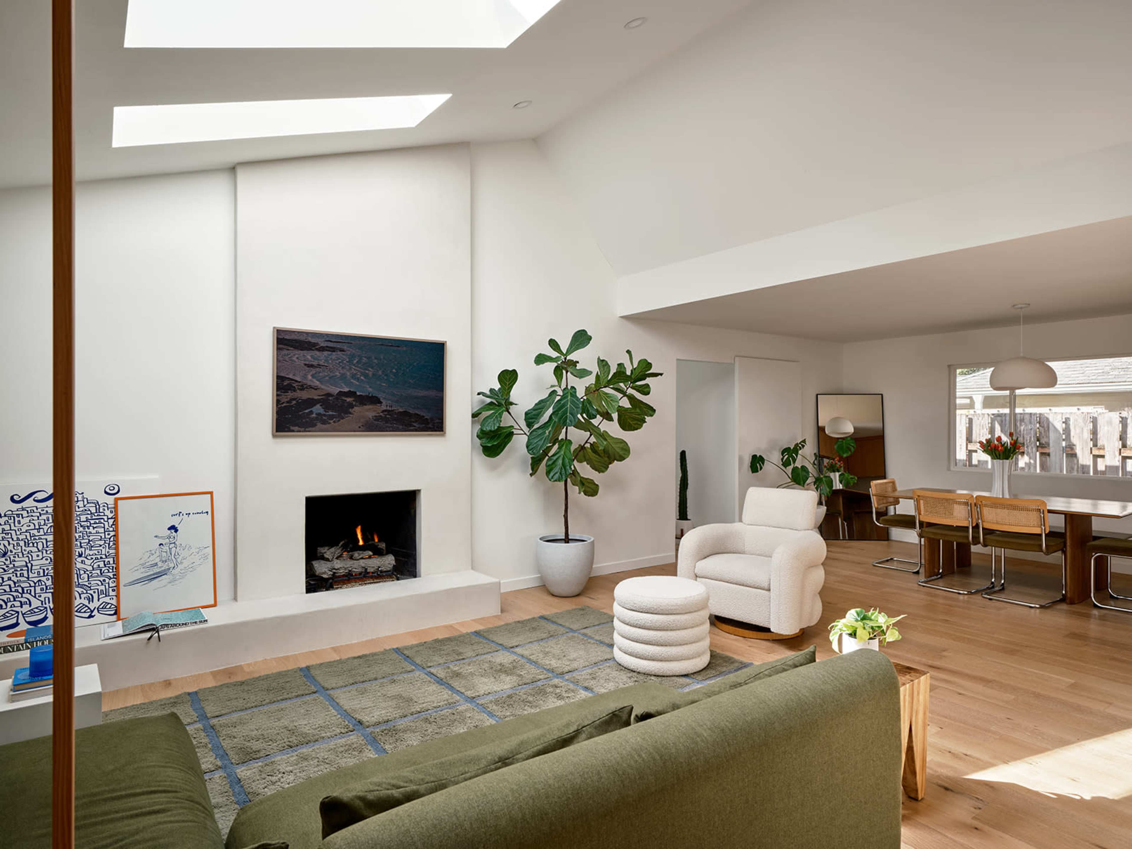 The image shows a modern living room with a skylight, a fireplace, a large plant, and a dining area in the background.