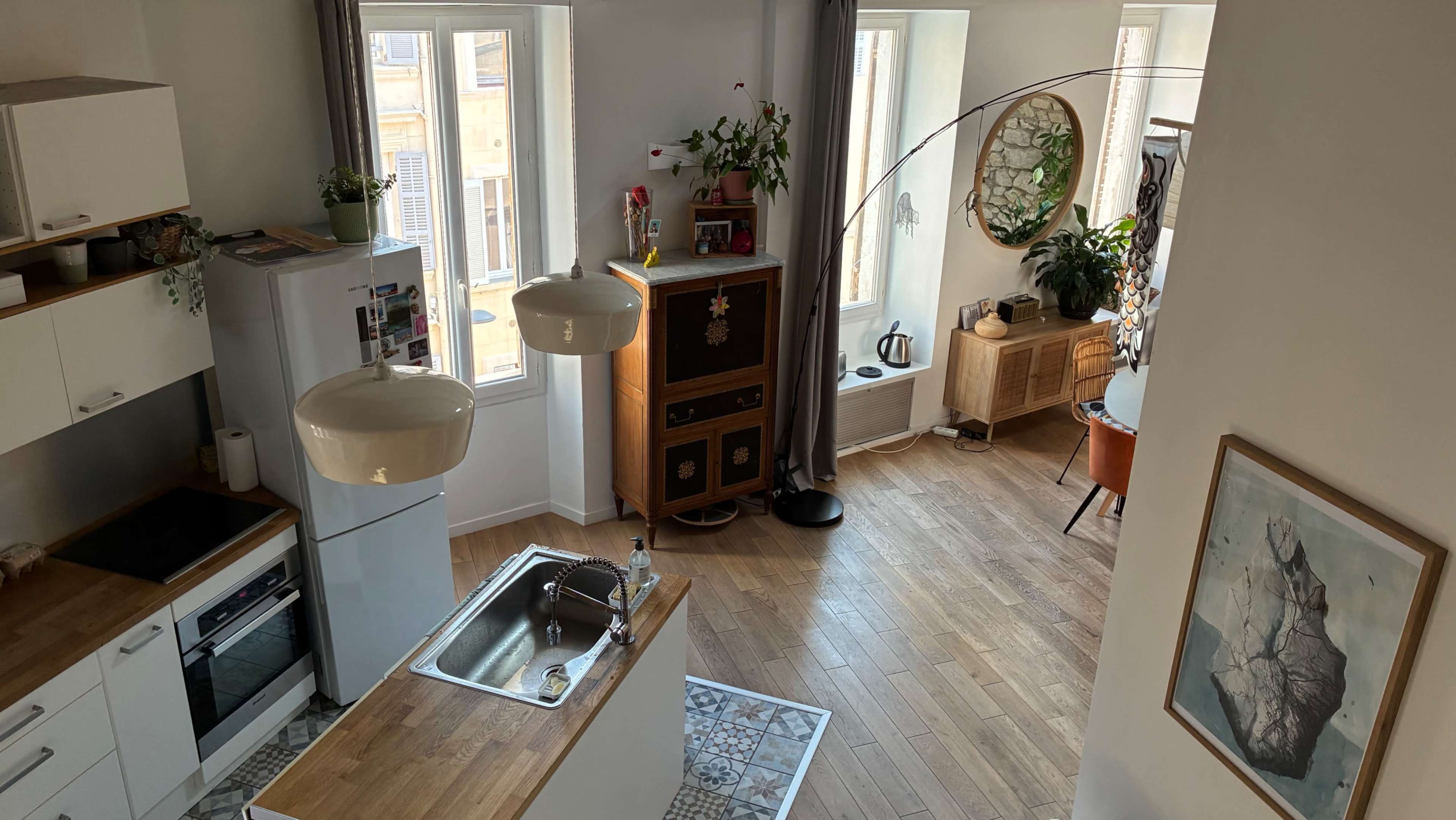 The image shows a bright, modern living space with a kitchen area on the left and a cozy seating area with plants and decorative furniture on the right.