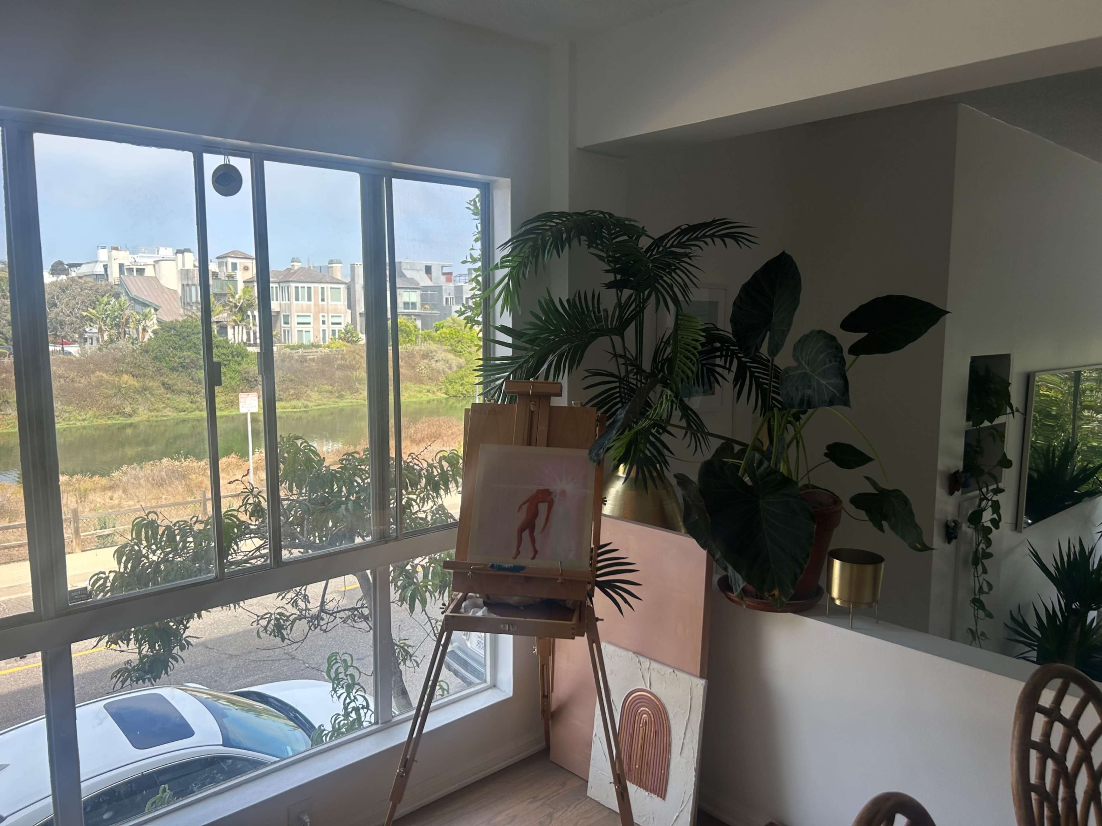 An interior space with a large window overlooking a body of water, an artist's easel displaying a painting, and several potted plants near a mirror.