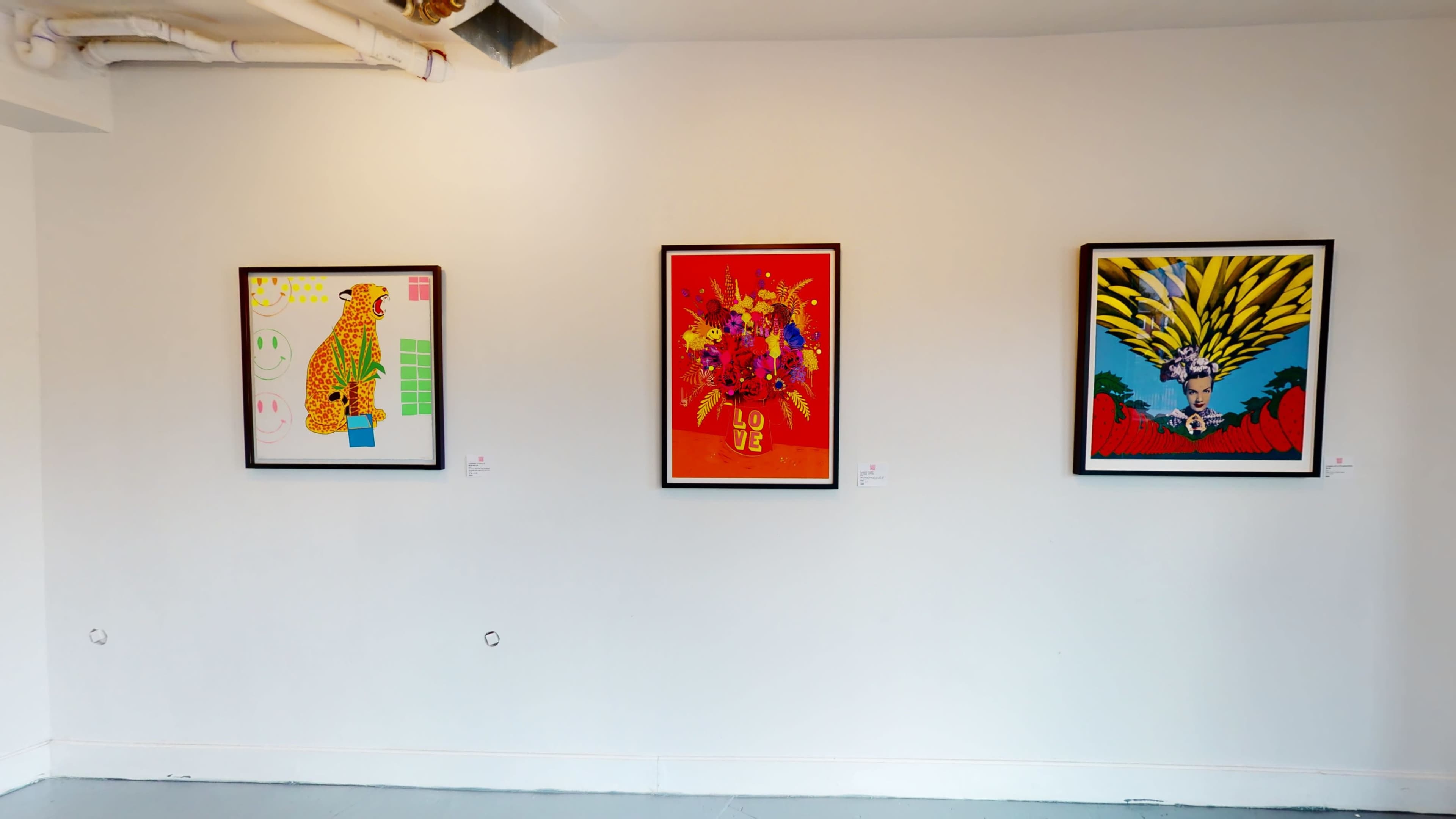 Three framed artworks hang on a white wall in an art gallery, featuring vibrant colors and abstract designs.